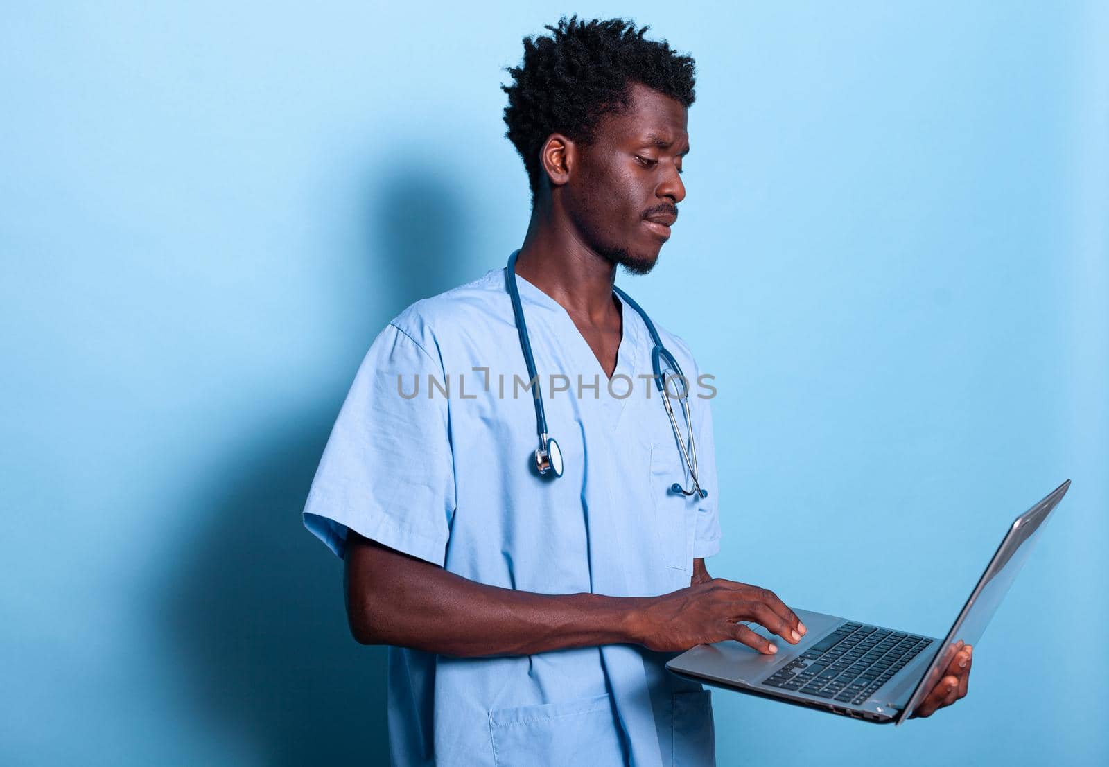 African american man nurse looking at laptop in hand. Black medical assistant with blue uniform and stethoscope holding modern device, standing over isolated background. Healthcare specialist