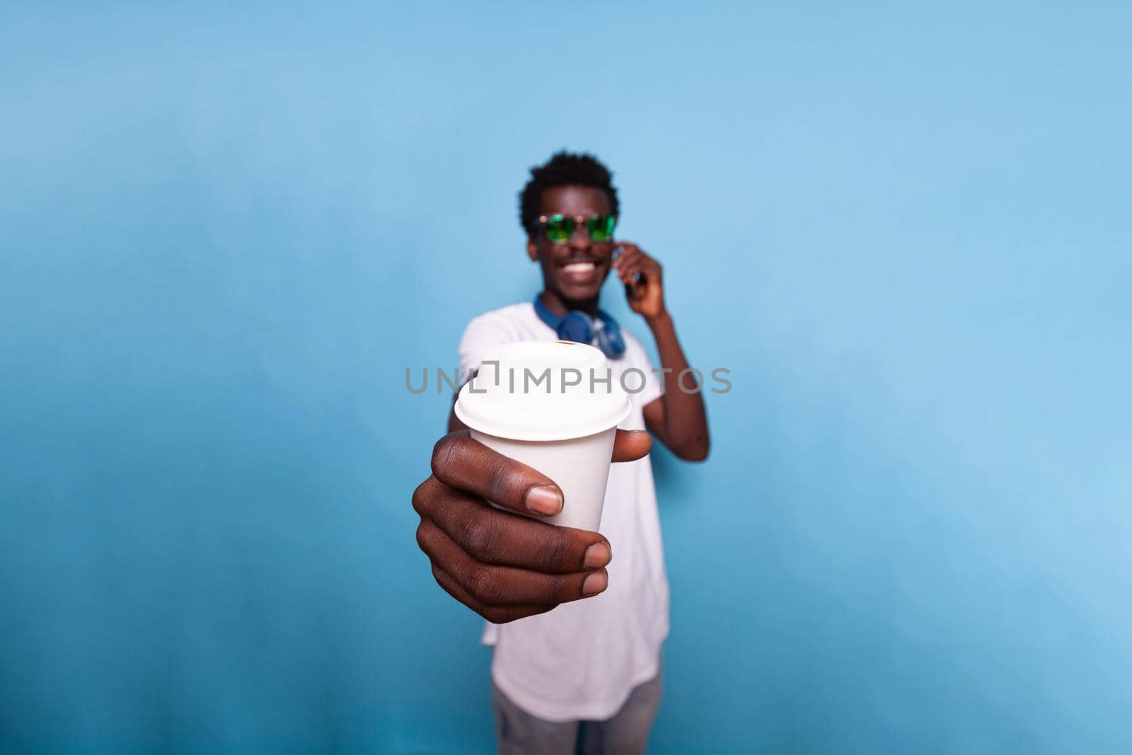 Carefree man holding coffee drink close to camera in studio. Casual adult talking on smartphone call and wearing sunglasses while showing beverage over isolated background. Person smiling