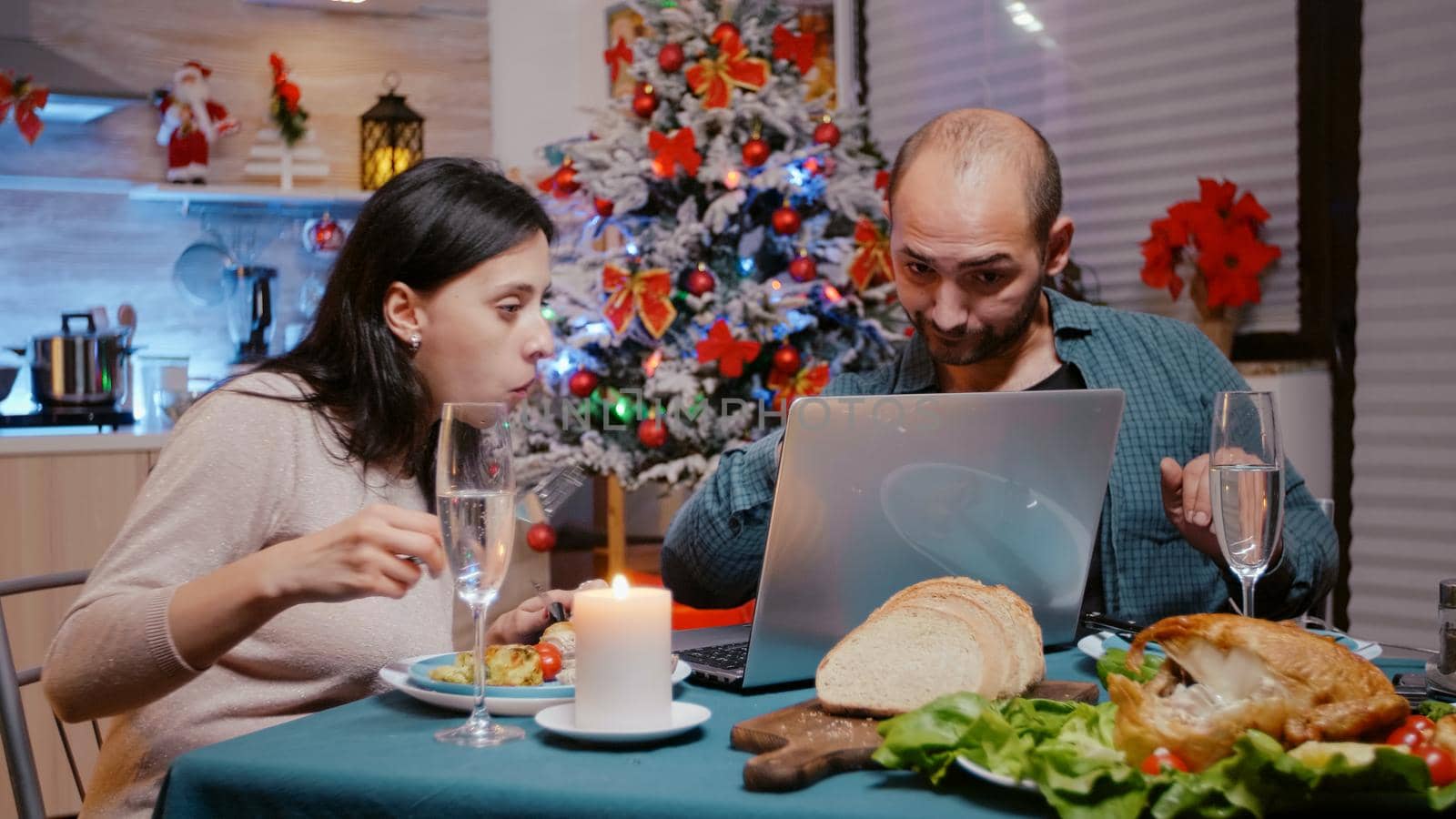 Festive couple looking at laptop and enjoying christmas dinner with technology. People eating chicken and drinking champagne while using device. Man and woman celebrating holiday.