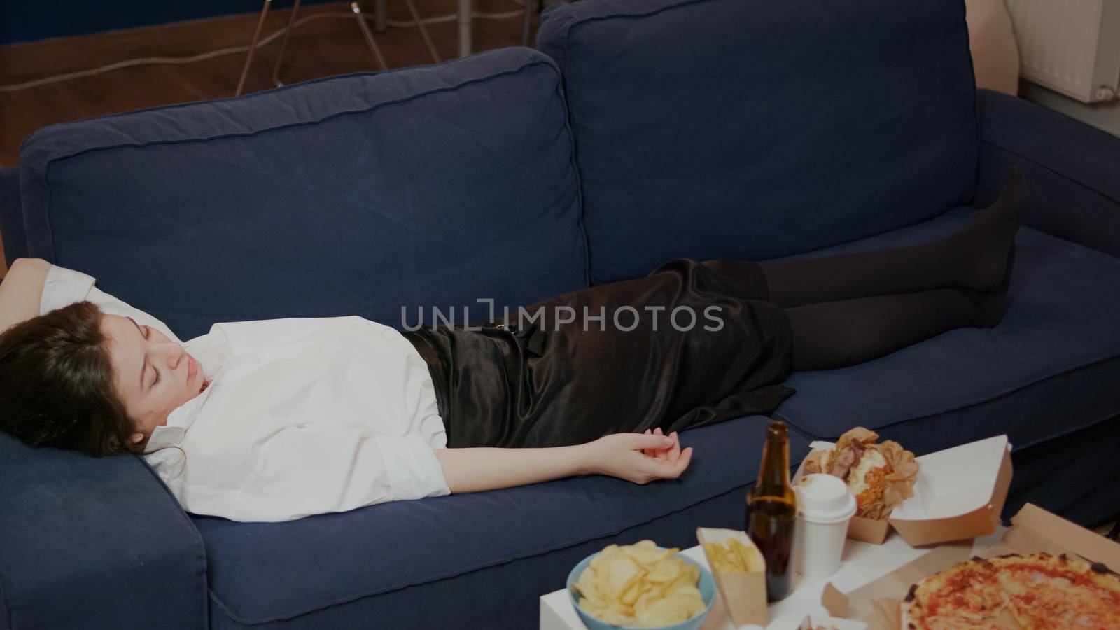 Young person falling asleep on couch dropping TV remote at home. Tired woman from work sleeping after eating takeaway food in living room. Adult taking nap while watching television