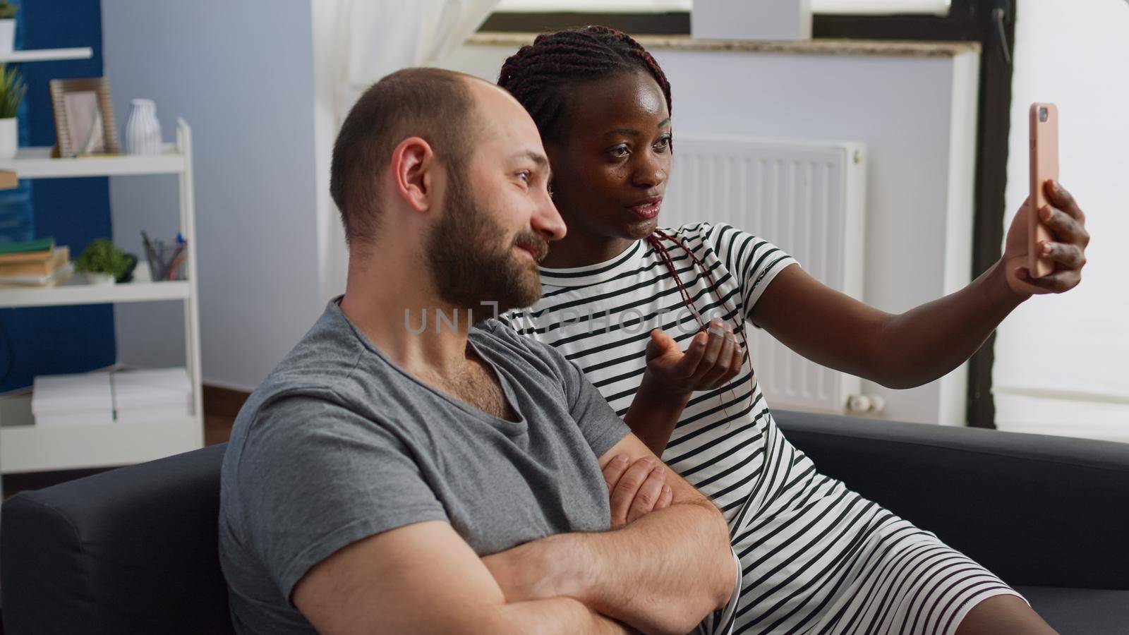 Interracial couple talking and waving on video call conference using modern smartphone, sitting on couch. Multi ethnic people with technology chatting using online remote communication