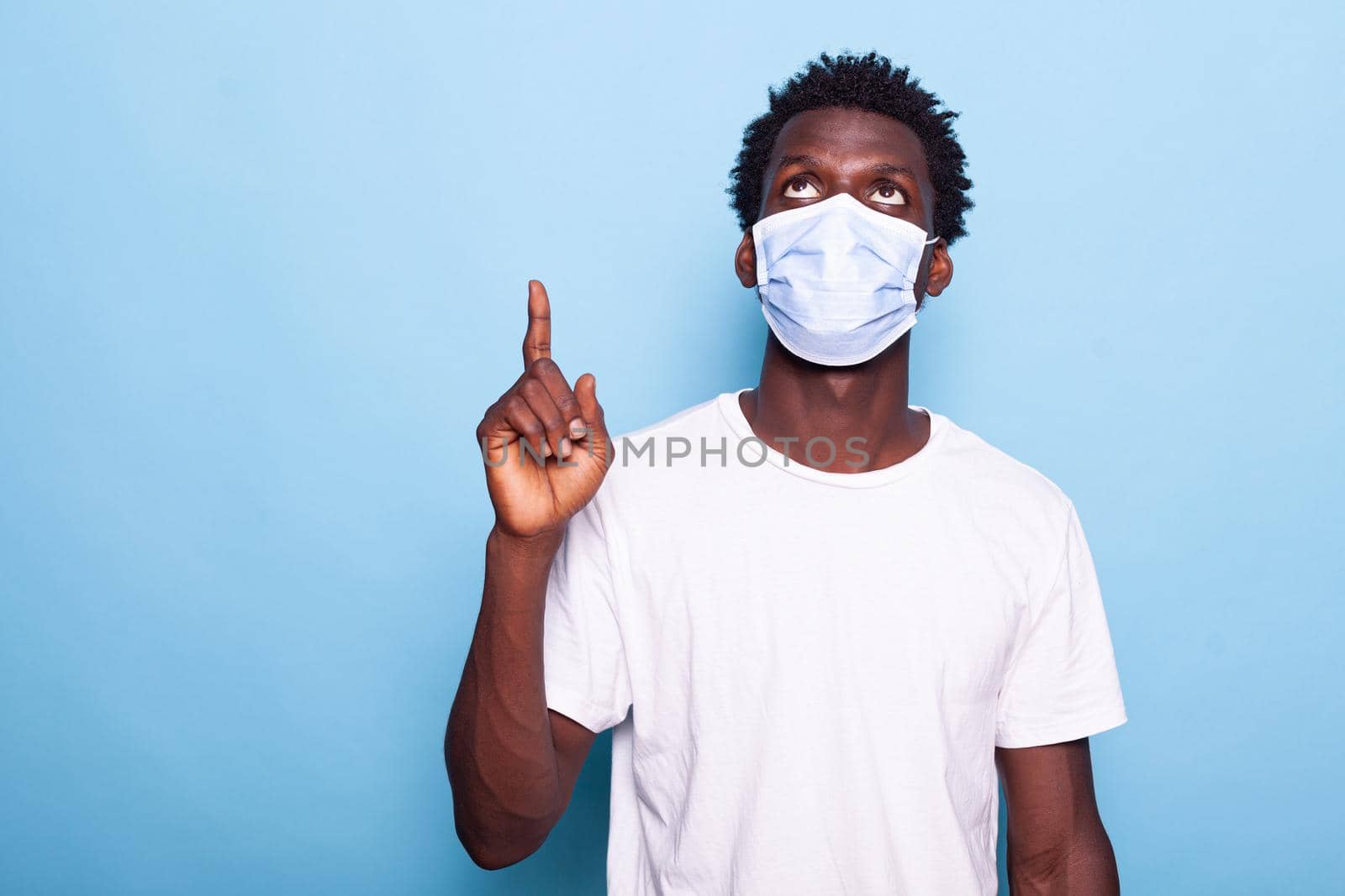 Casual man with face mask against coronavirus pointing up with index finger. Young adult wearing protection for virus pandemic while indicating direction, doing sign with hand.