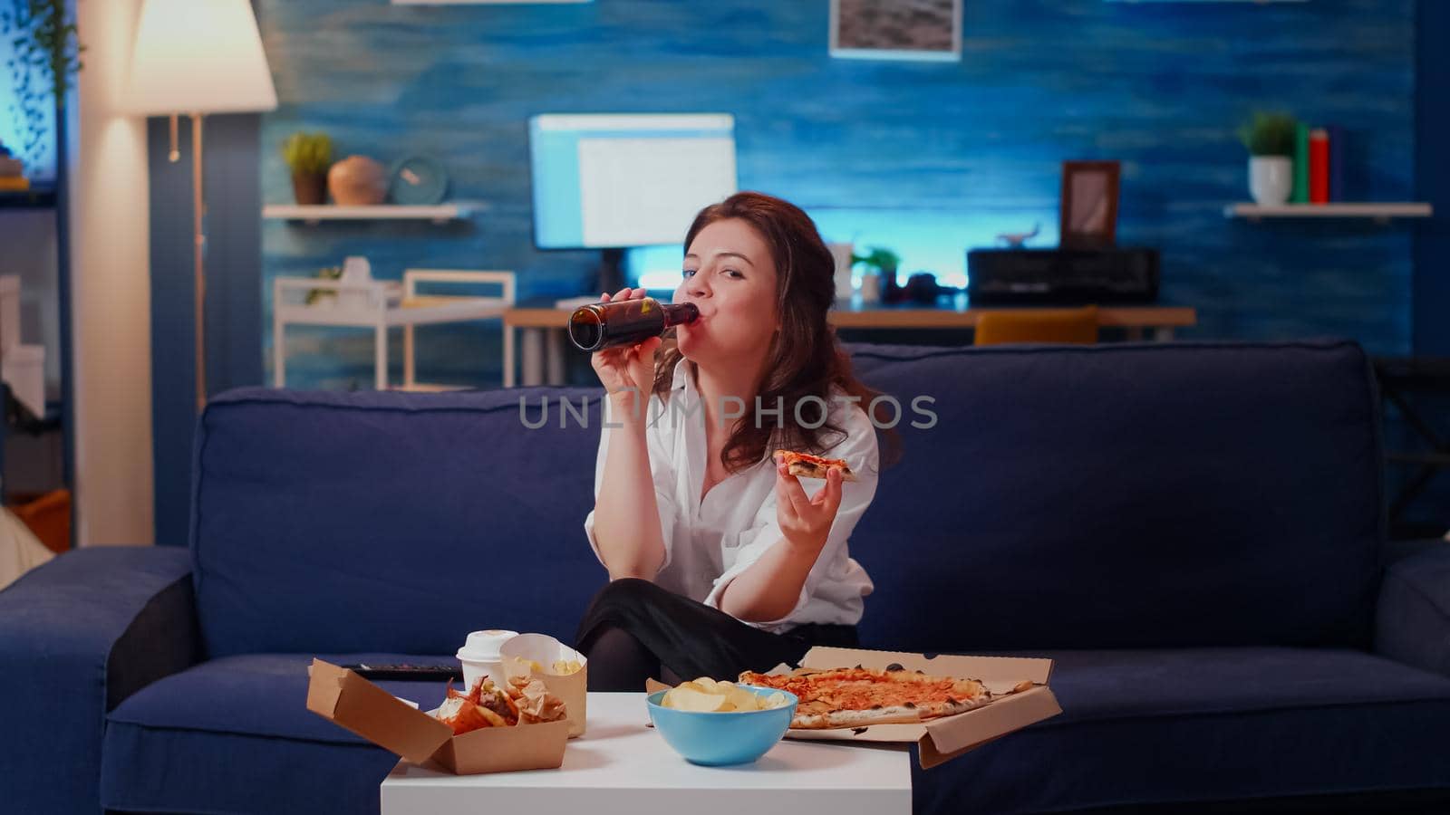 Person eating slice of pizza on couch and looking at camera in living room. Caucasian woman with fast food meal from delivery at home while watching television and laughing after work