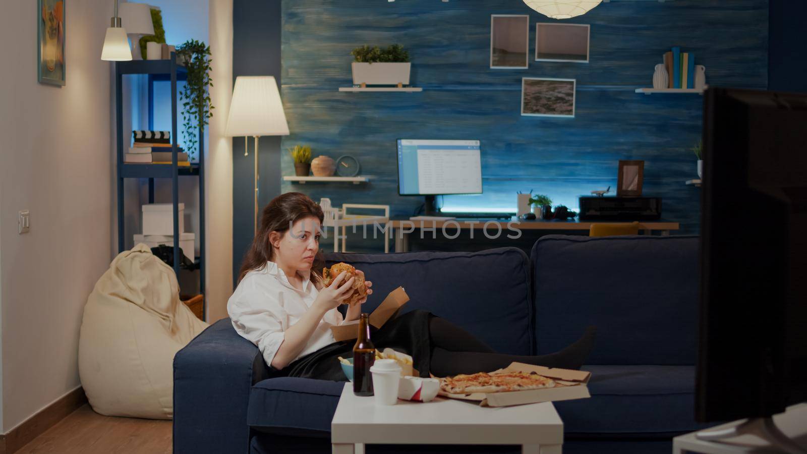 Young woman laying on couch eating burger and laughing watching comedy movie on television. Adult enjoying takeaway meal with hamburger and beer while relaxing on sofa looking at TV