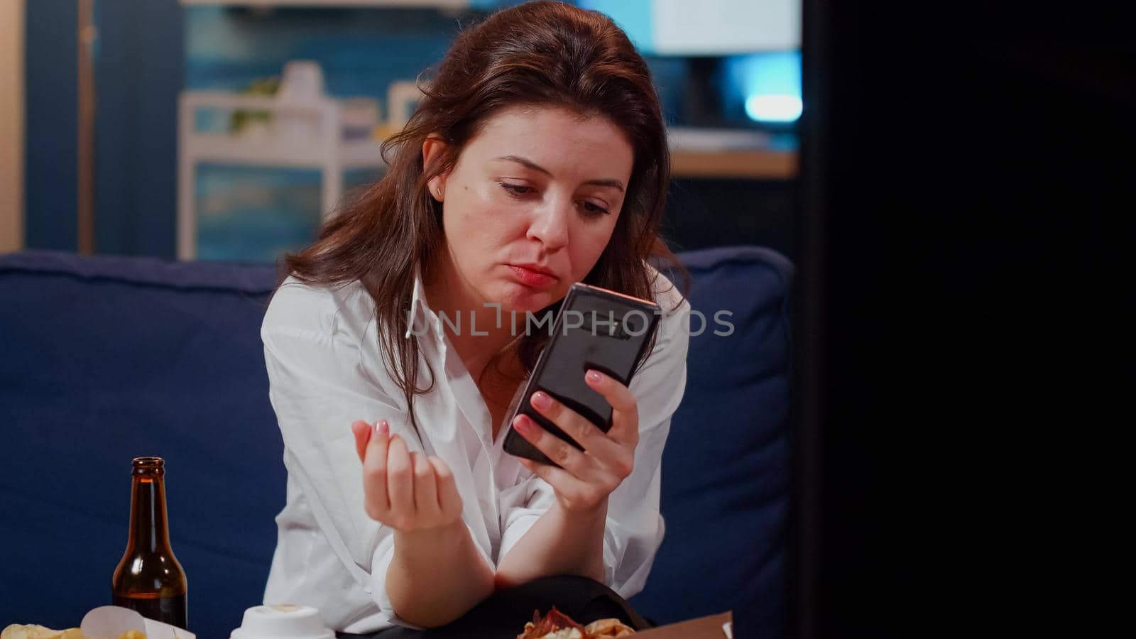 Caucasian woman biting slice of pizza and holding smartphone in living room. Young adult using technology while watching TV and enjoying fast food meal and bottle of beer from delivery