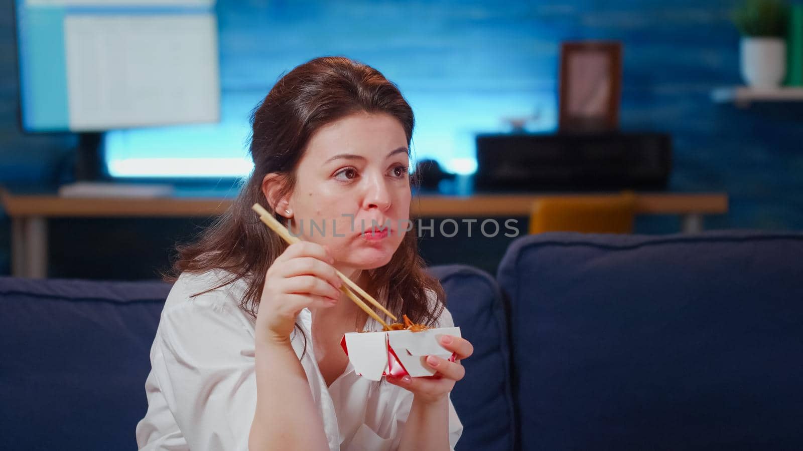 Close up of business woman eating asian food at TV after work on couch. Young person using chopsticks for takeout chinese meal with noodles while looking at television in living room