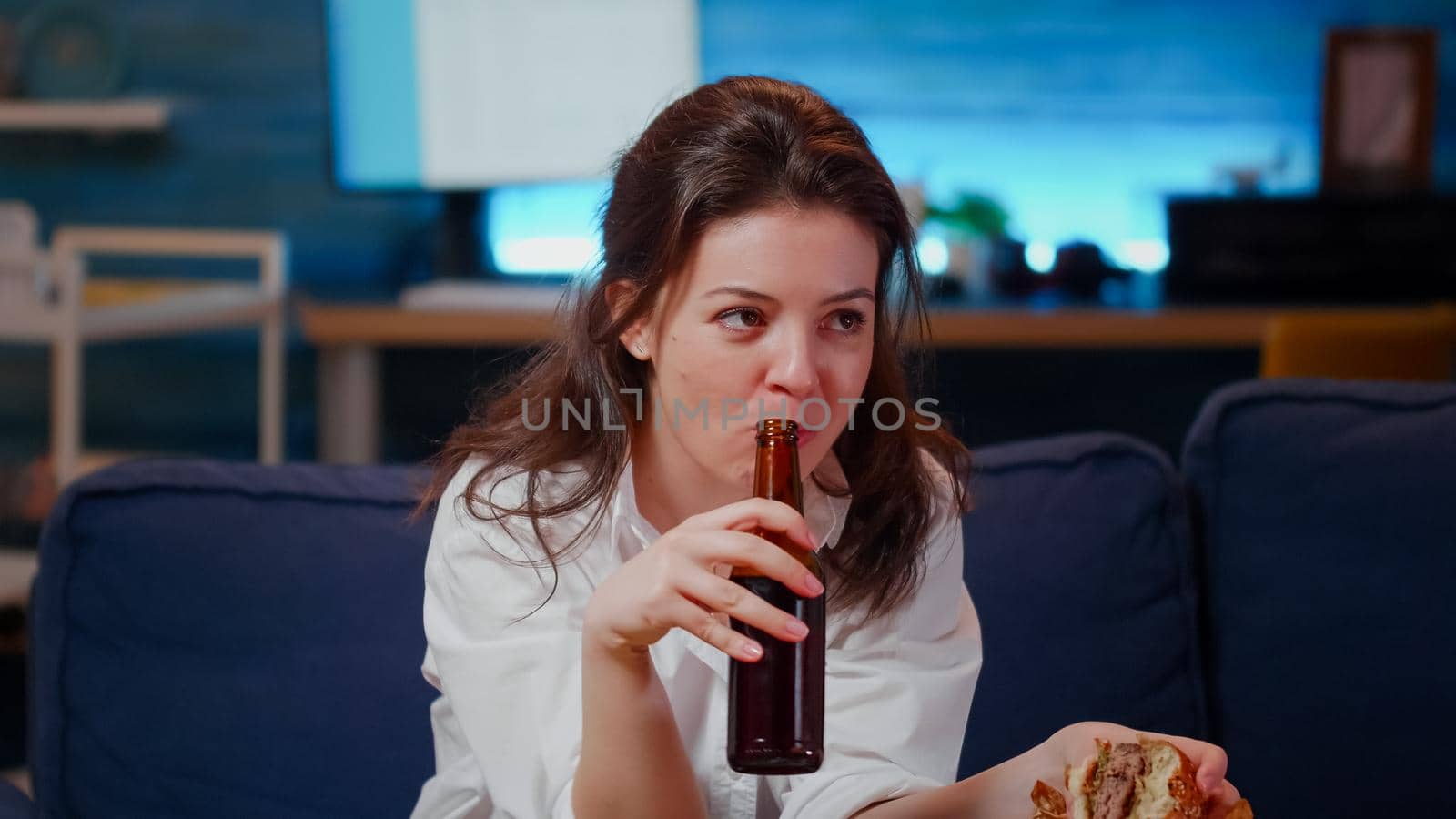 Young person with fast food meal watching television sitting on sofa after work. Caucasian woman eating hamburger and fries while drinking beer from bottle in living room at home.