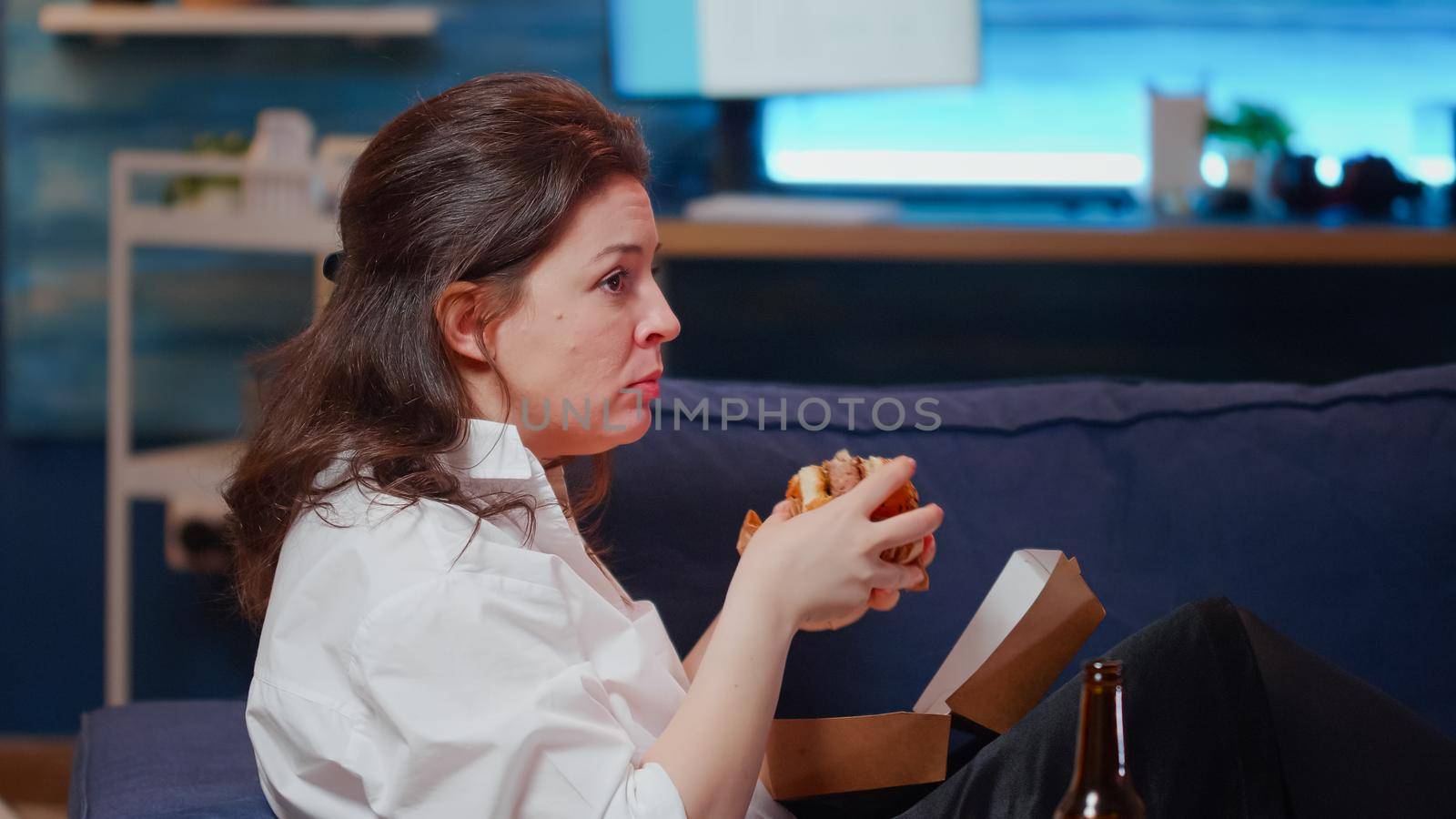 Caucasian person enjoying free time with fast food and television relaxing on couch in living room. Woman eating burger for dinner after work while laying on sofa with TV and takeaway meal