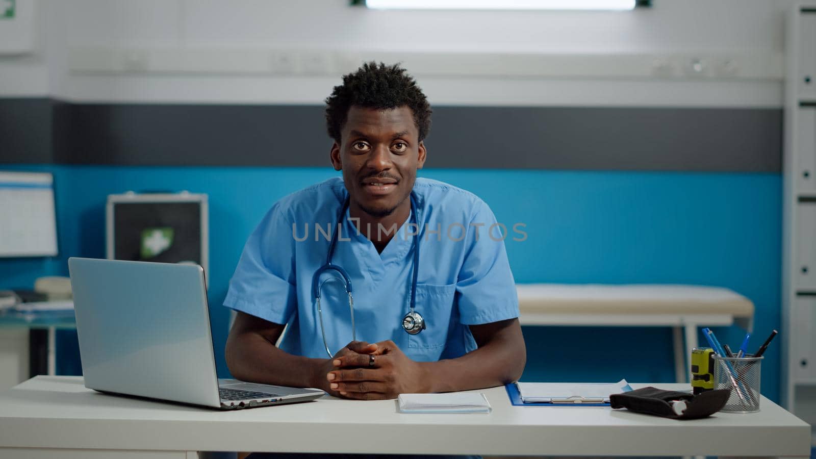 Close up of black medical assistant smiling sitting at desk in healthcare office. African american nurse looking at camera in cabinet with technology, documents and professional equipment