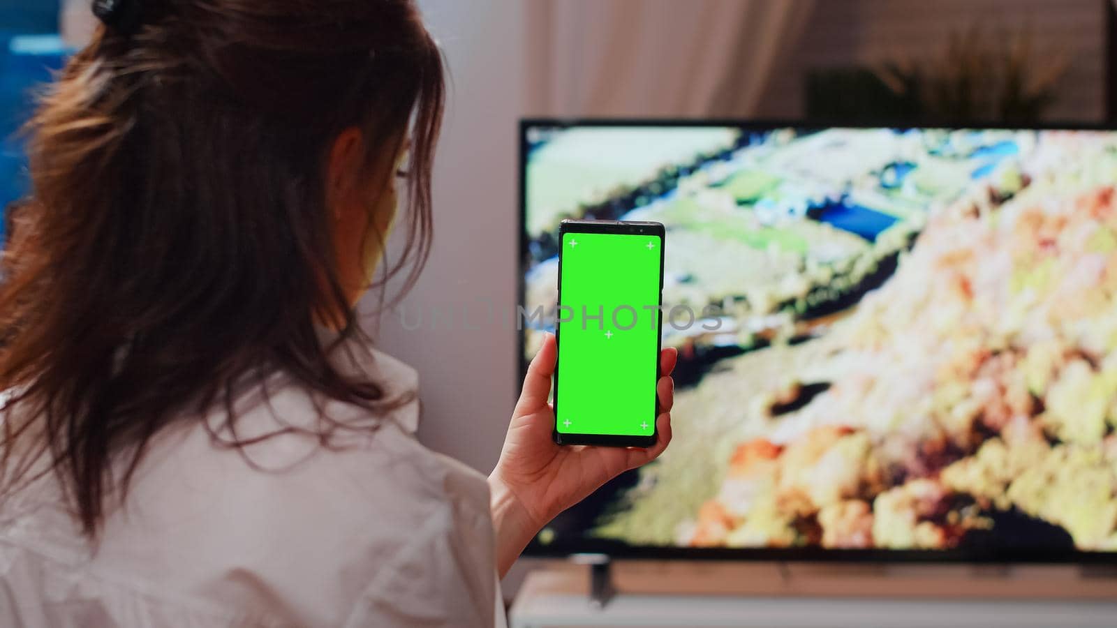 Caucasian person vertically holding green screen on smartphone in living room. Young woman looking at chroma key display with mockup template and isolated background on modern gadget