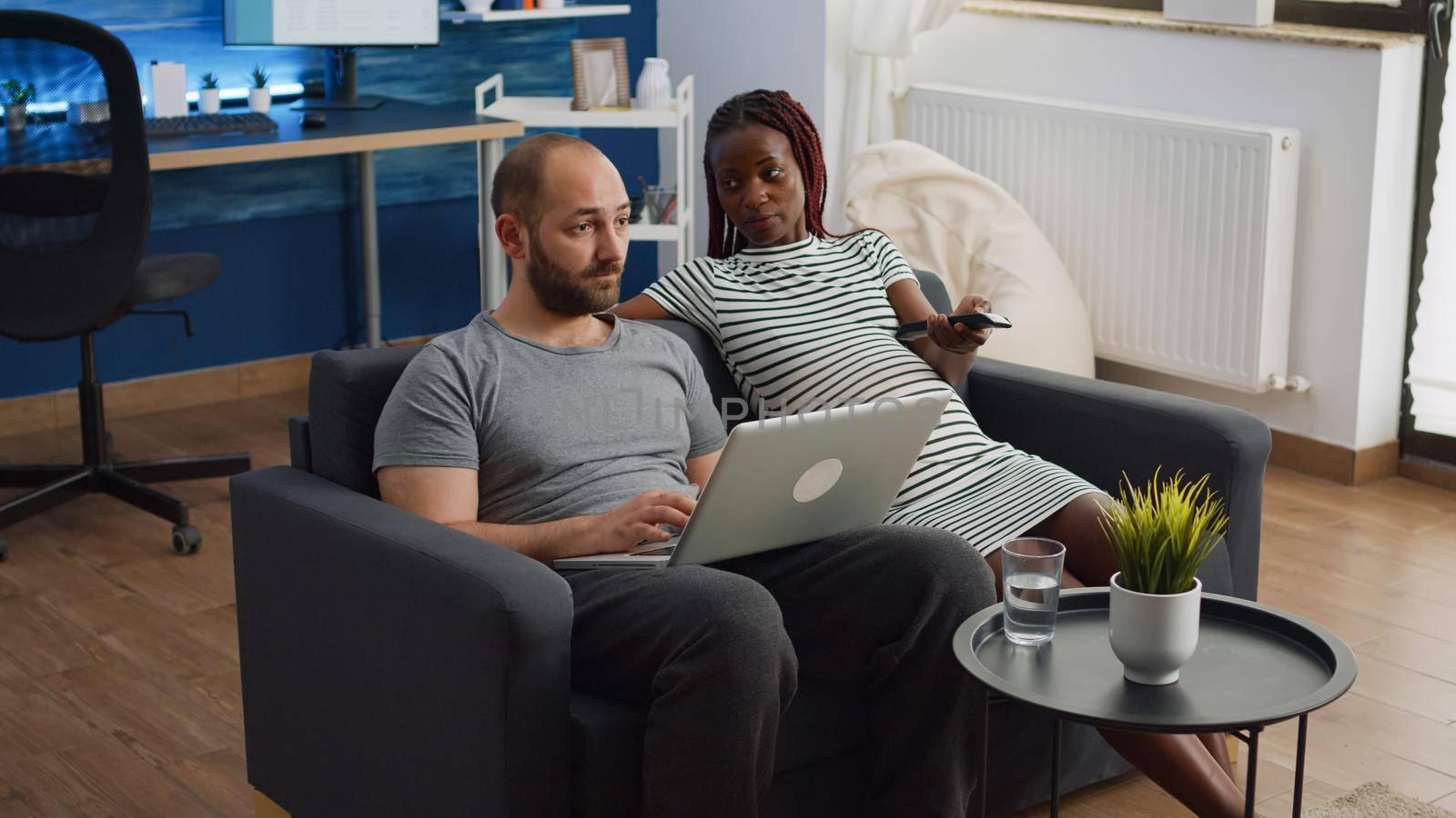 Interracial couple with pregnancy relaxing together at home. Pregnant black woman switching channels with TV remote control and caucasian man using laptop while sitting in living room