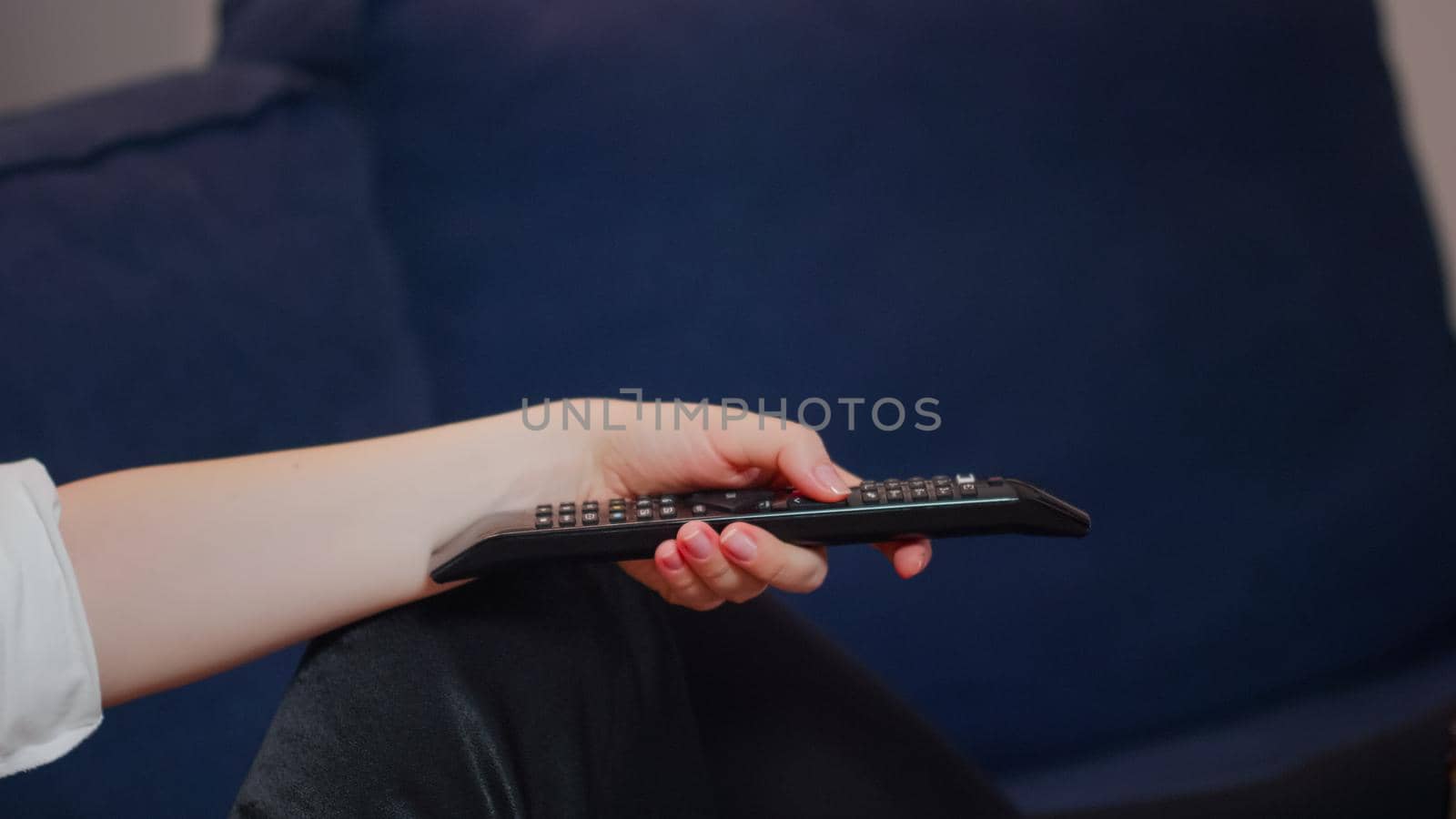 Close up of hand holding remote for television while laying on couch. Caucasian person switching channels relaxing on living room sofa. Young adult watching TV after work at home