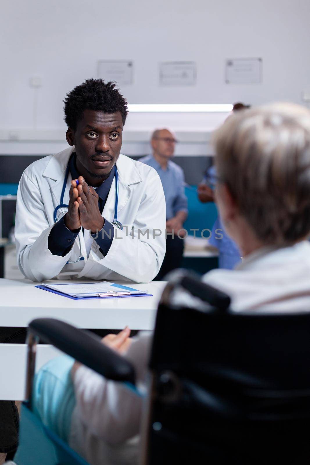 Young black doctor talking to invalid patient at office desk for healthcare consultation. African american medic meeting senior woman sitting in wheelchair discussing disease and treatment