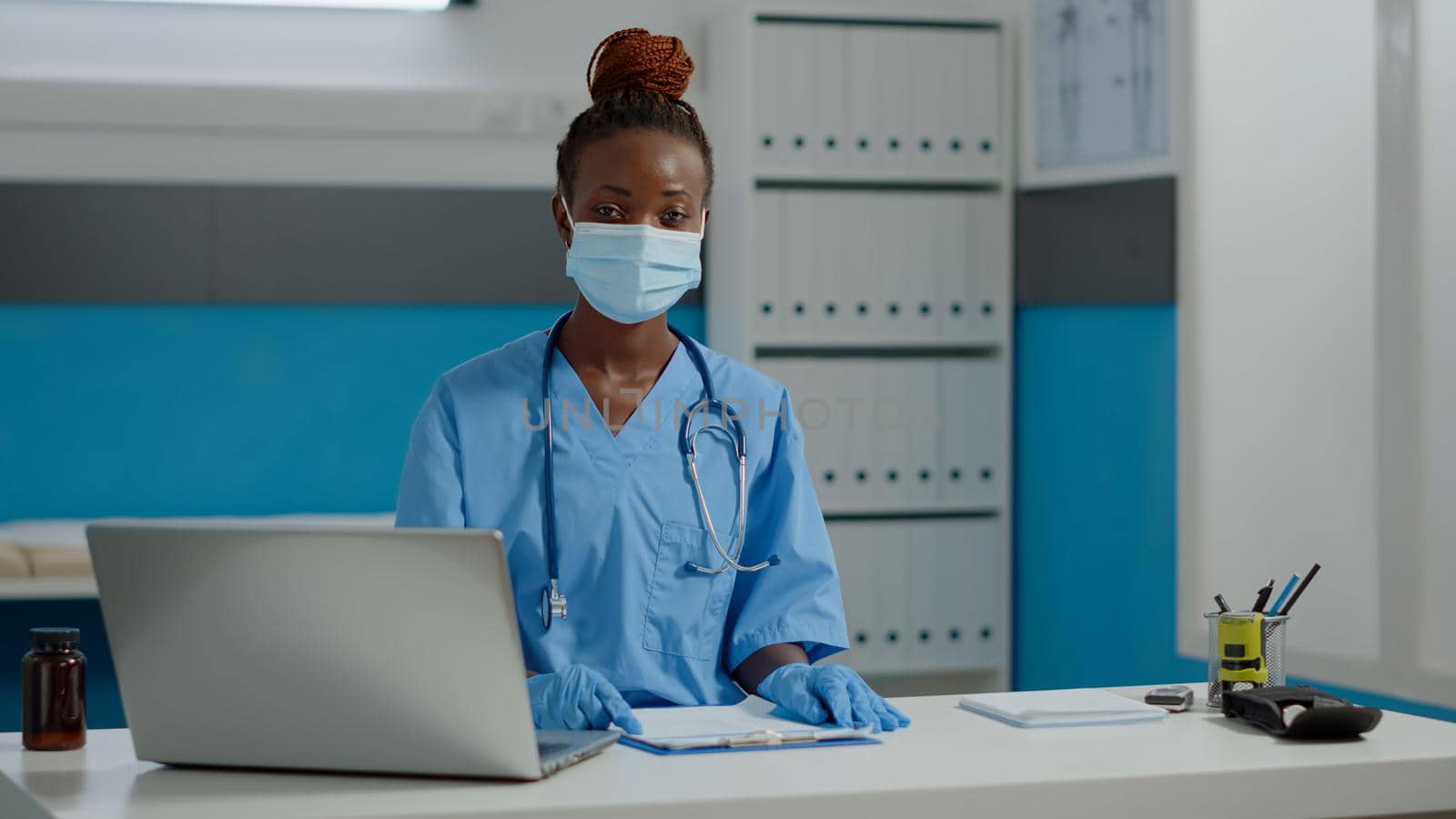 Portrait of nurse sitting at desk with laptop and documents while wearing uniform, gloves and face mask at medical facility. Assistant looking at camera with bottle of pills on table