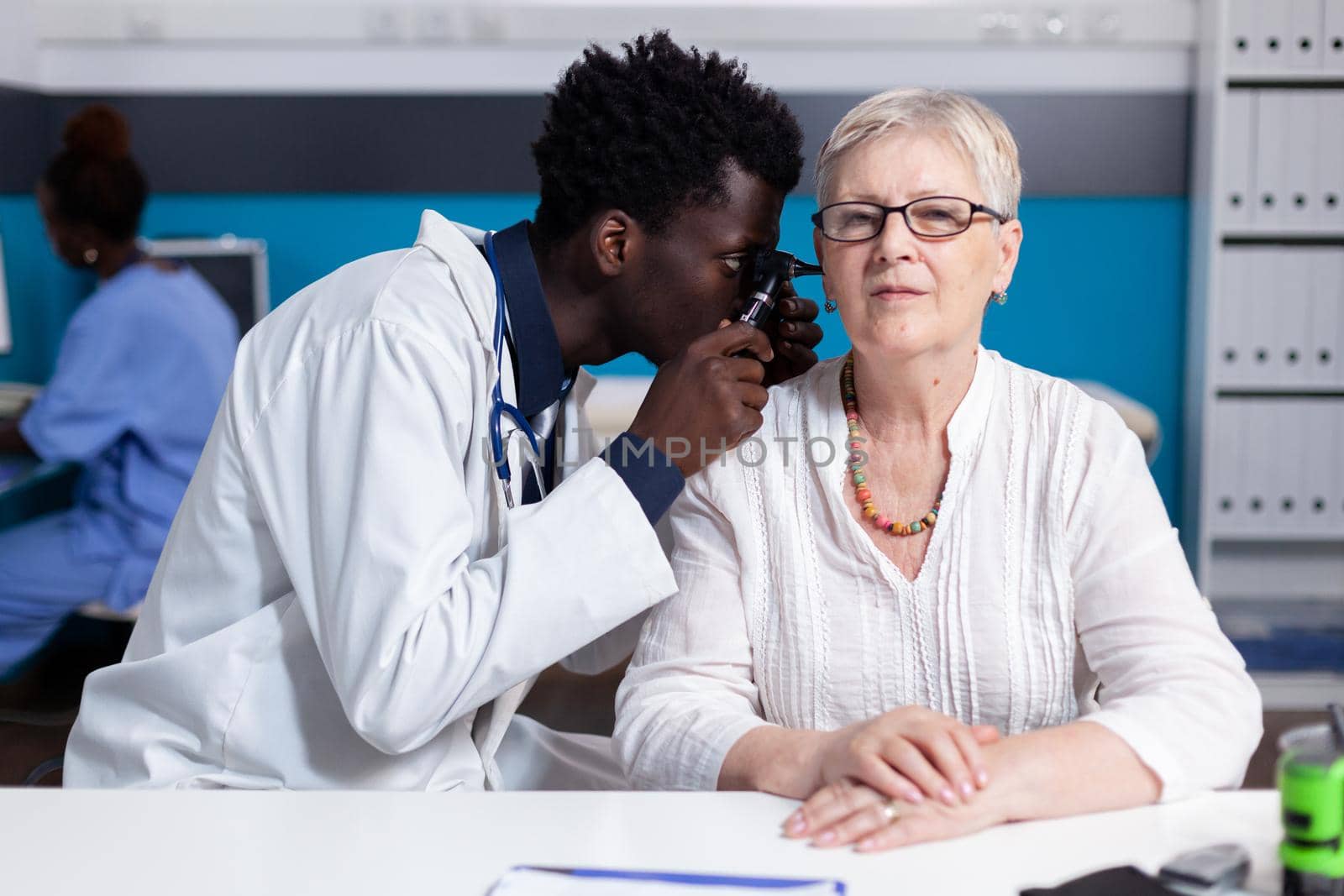 Black young doctor using otoscope on elder patient for ear consultation and examination. African american medic with metal tool for senior woman with healthcare problems in cabinet