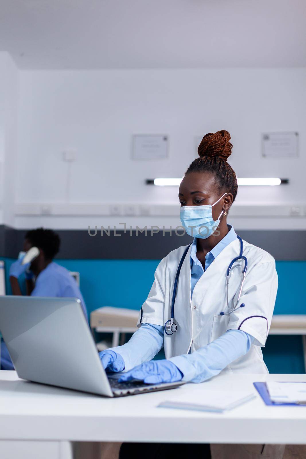 Woman of african ethnicity working as doctor in medical cabinet at desk. Black medic with stethoscope, uniform and face mask using modern laptop technology while sitting in office