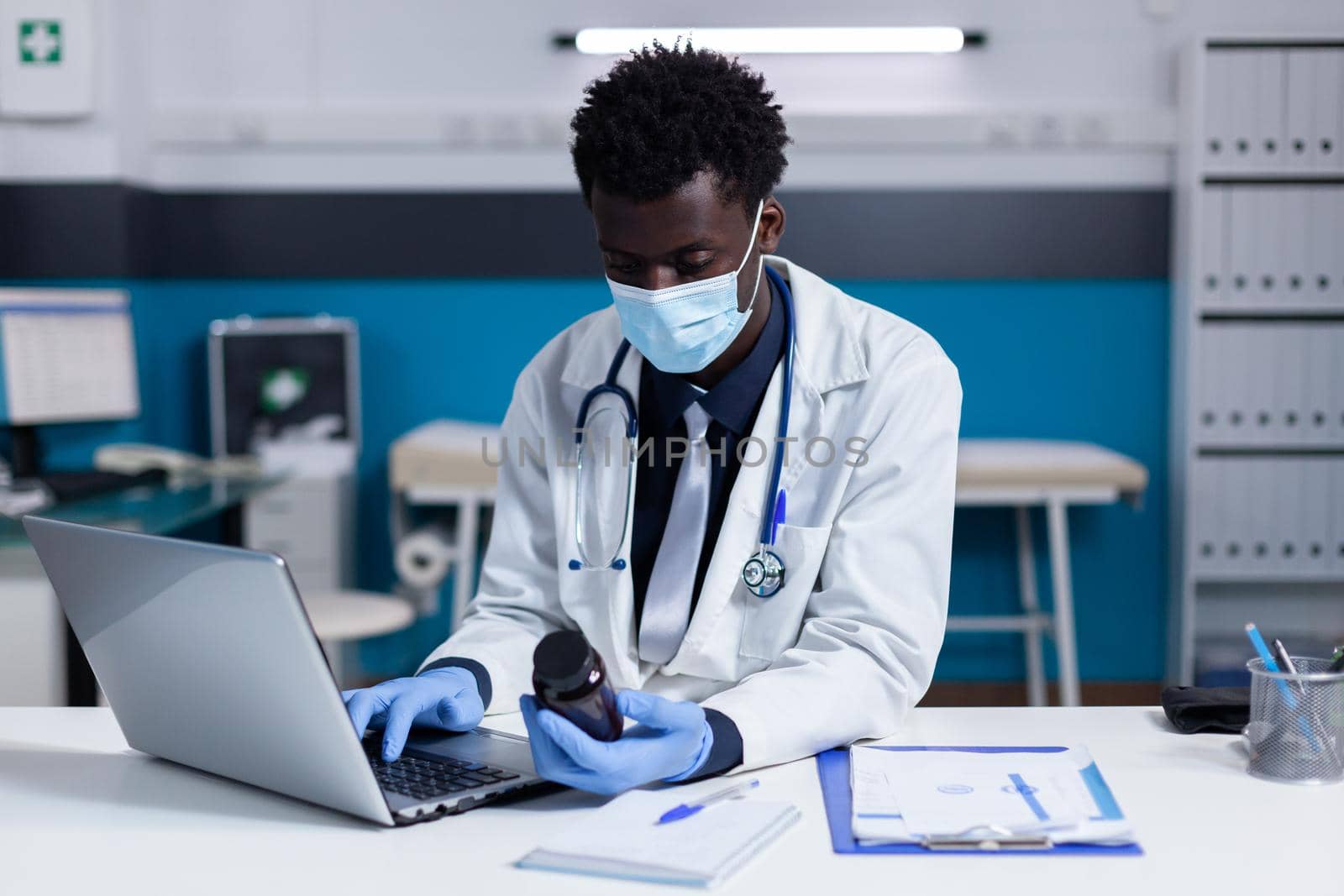 Black person with medic profession using laptop on desk holding bottle of medicament in medical cabinet. African american professional doctor typing prescription on keyboard while sitting