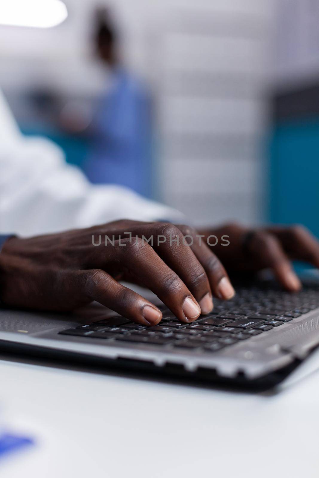 Close up of black hands typing on laptop keyboard while sitting at white desk in medical office. African american man wearing medical coat using computer device and modern technology