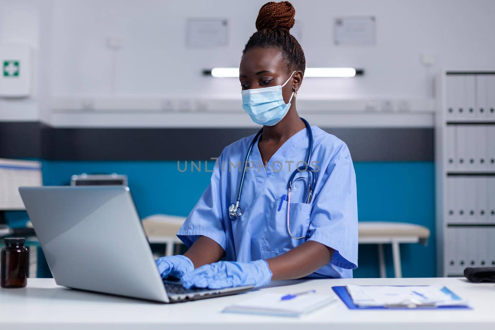 Black woman with nurse profession using modern laptop and technology while sitting at desk in medical cabinet. African american specialist with stethoscope wearing face mask for protection