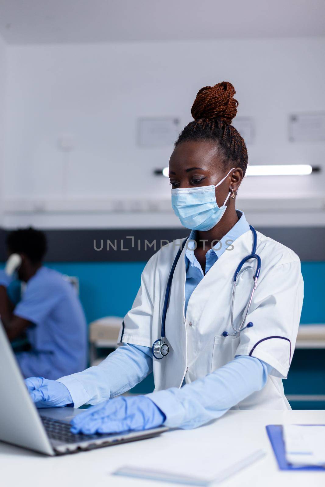 Woman of african ethnicity working as doctor in medical cabinet at desk. Black medic with stethoscope, uniform and face mask using modern laptop technology while sitting in office