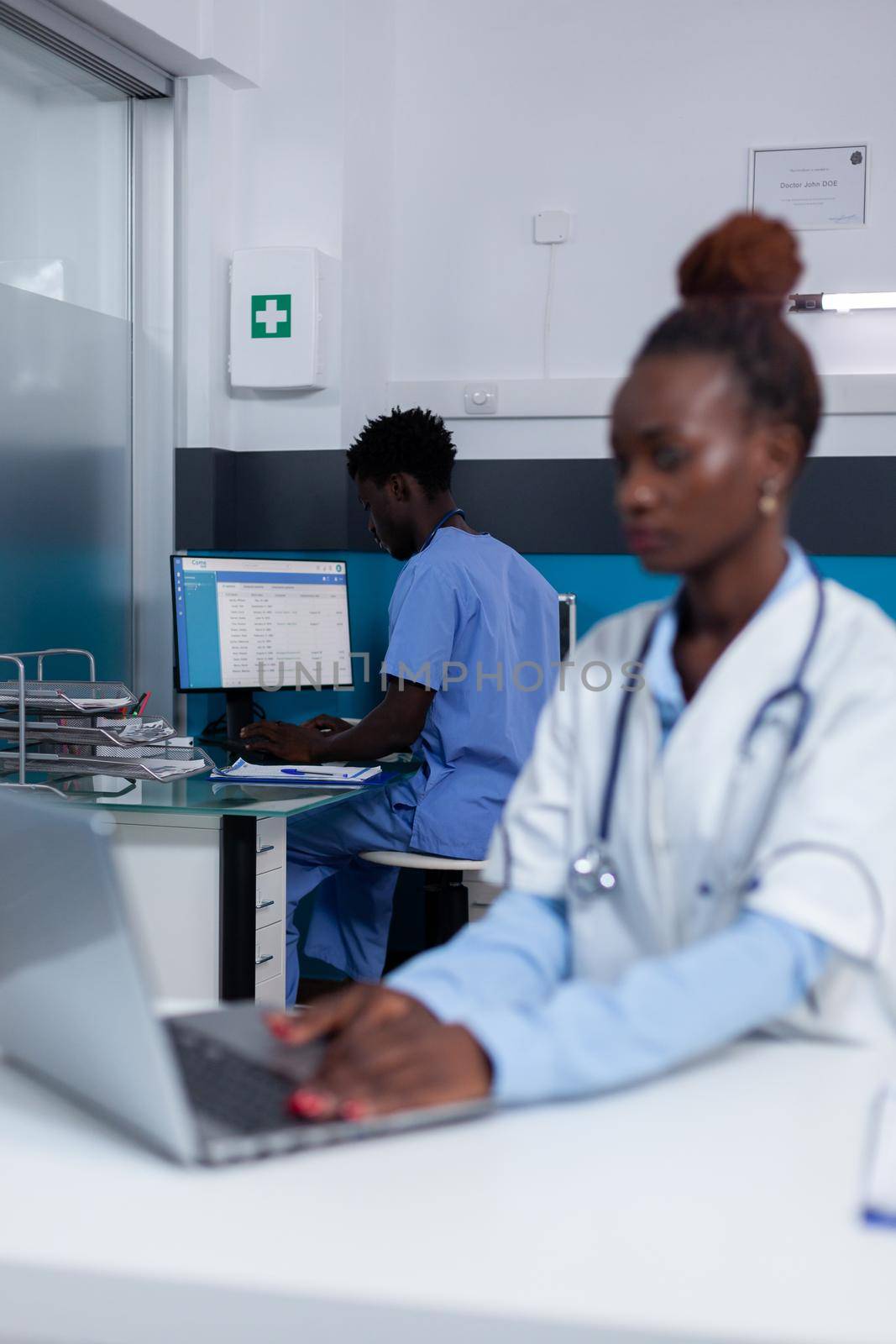 Portrait of african american woman with doctor profession sitting at desk while using laptop and technology in healthcare office. Black medic with modern device and man nurse in background