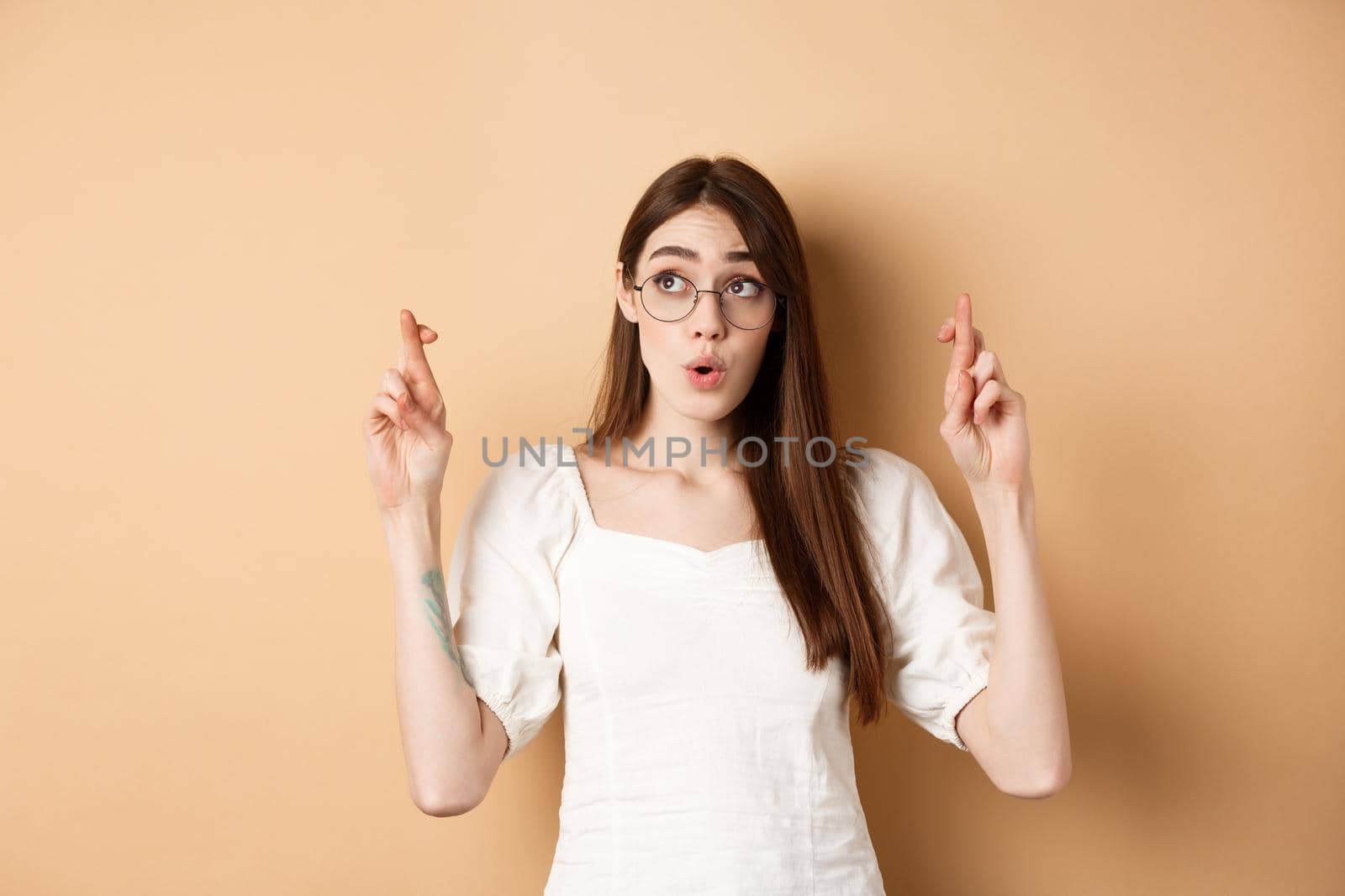 Excited girl hope dream come true, making wish with fingers crossed, looking aside and praying, standing on beige background.
