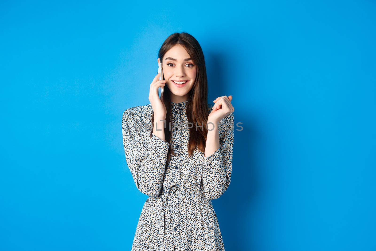 Excited woman receive good news on mobile phone, smiling happy, talking on smartphone, standing on blue background.