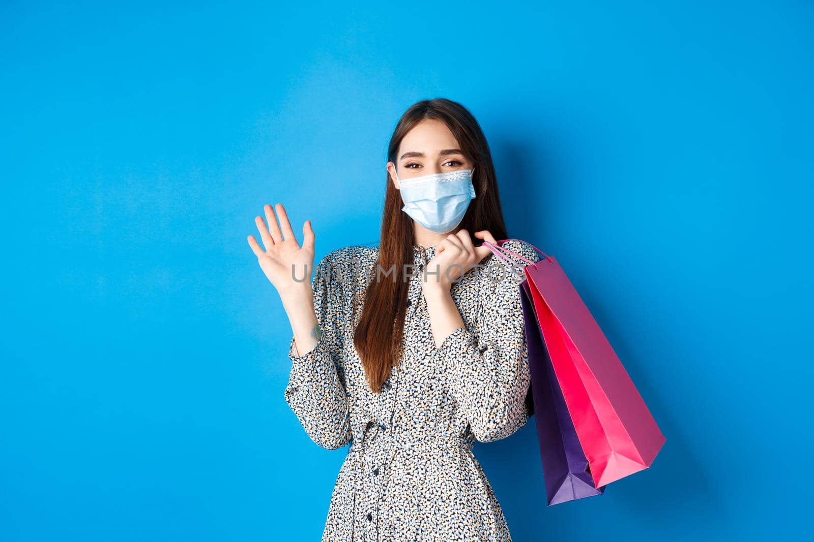 Covid-19, pandemic and lifestyle concept. Friendly cute woman wear medical mask in shopping mall, waving hand and holding bags, say hello, standing on blue background.