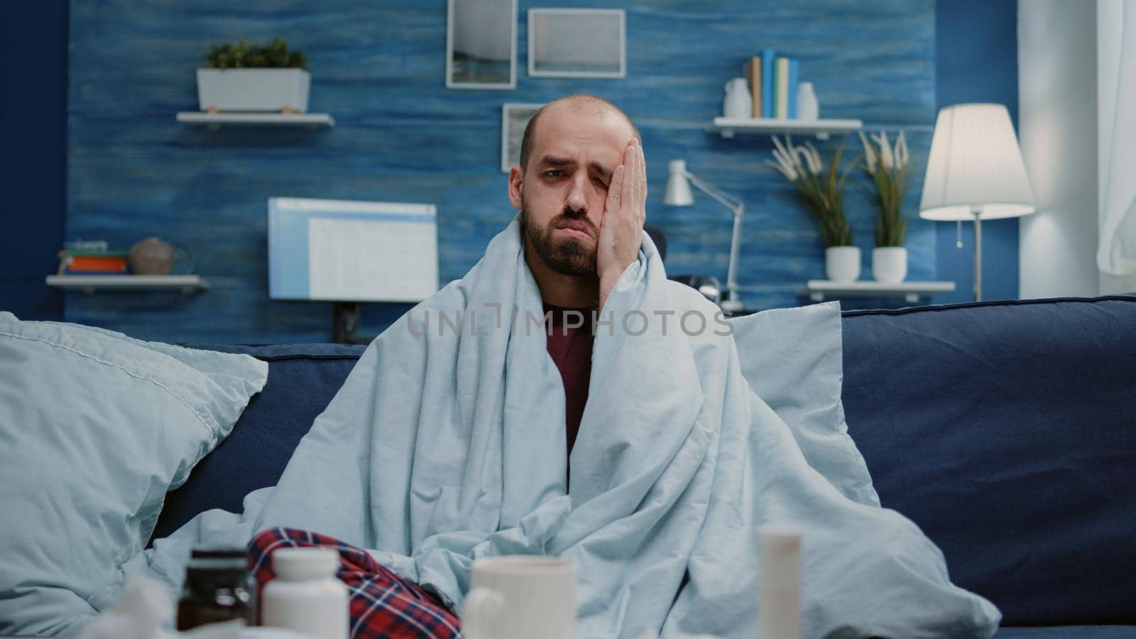 Close up of ill man with headache rubbing temples while looking at camera. Portrait of sick adult with coronavirus symptoms and migraine while feeling cold with blanket. Person with flu