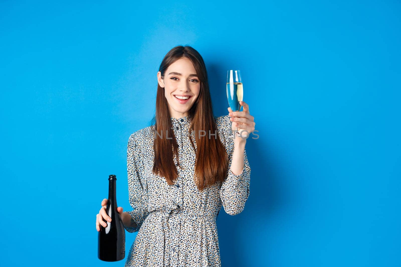 Celebration. Happy young woman in dress raising glass of champagne, holding bottle and smiling, having fun at party, standing on blue background.