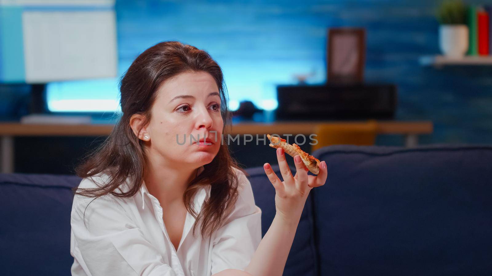 Young person eating slice of pizza while watching television on couch at home. Woman holding bottle with alcoholic beverage and enjoying takeaway fast food meal in living room after work