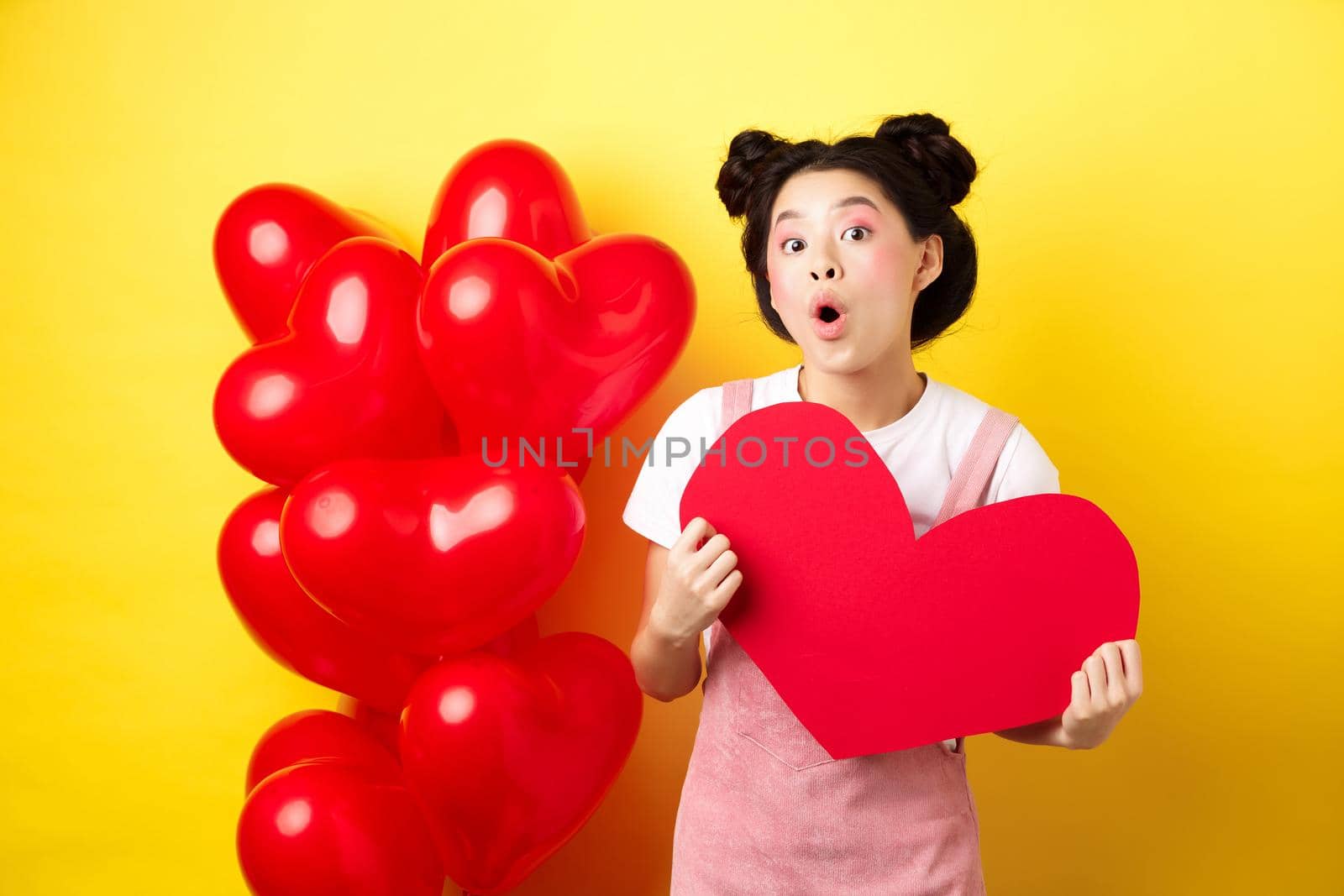Happy Valentines day. Silly asian girl showing big red heart postcard and looking amazed, standing near red romantic balloons, yellow background.