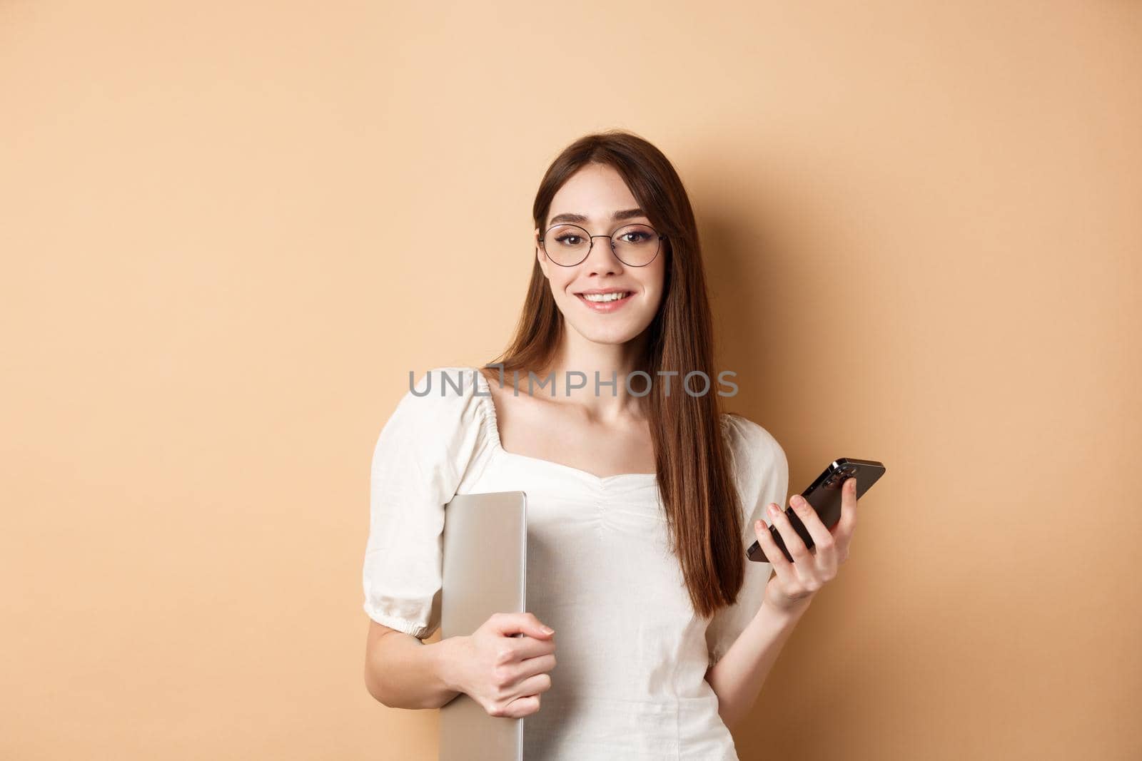 Elegant working woman in glasses holding laptop computer and smartphone, smiling pleased at camera, standing on beige background by Benzoix