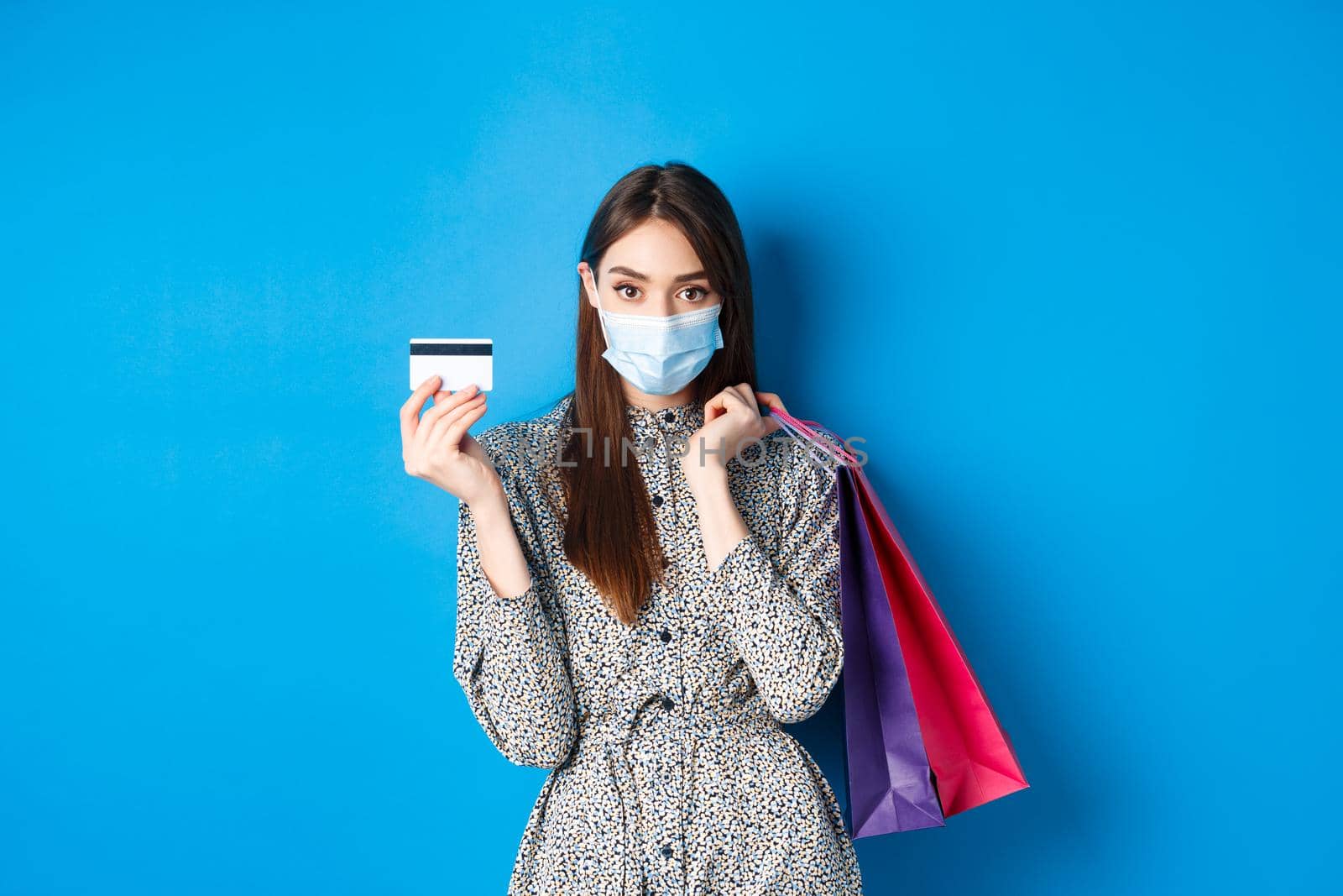 Covid-19, pandemic and lifestyle concept. Stylish woman wearing medical mask on shopping as preventive measure from coronavirus, showing plastic credit card and holding bags, blue background.