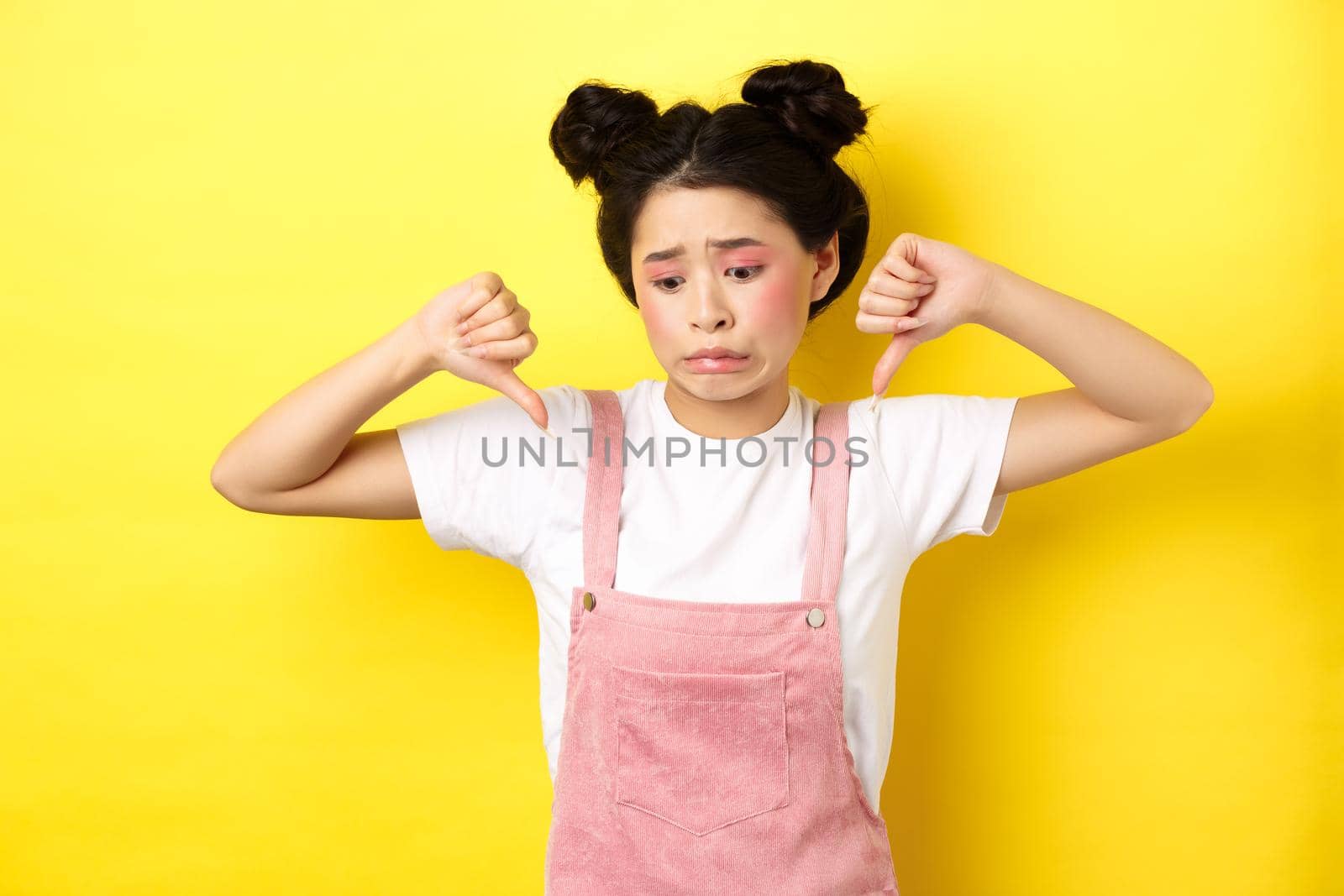 Asian beauty girl cringe from something disgusting, look away and showing thumbs-down to express dislike, negative emotions, standing in summer clothes on yellow background.