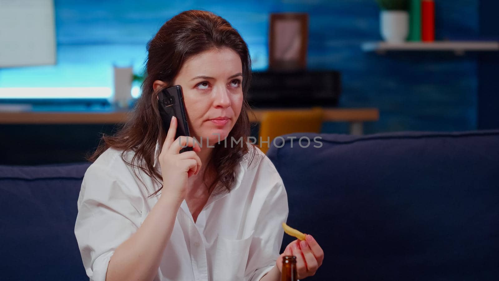 Annoyed woman talking on smartphone and eating at home on couch. Young adult using phone for business while trying to relax and enjoy takeaway meal after work, sitting in living room