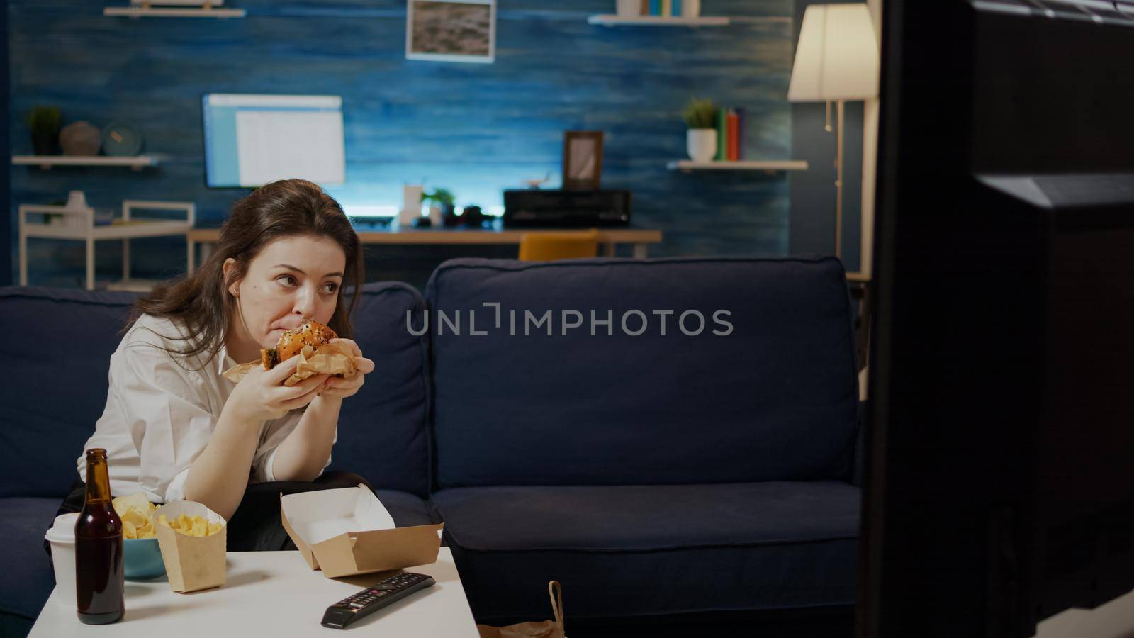 Caucasian woman eating hamburger from delivery bag while watching movie on television at home. Adult enjoying takeaway fast food meal and bottle of beer sitting on couch in living room