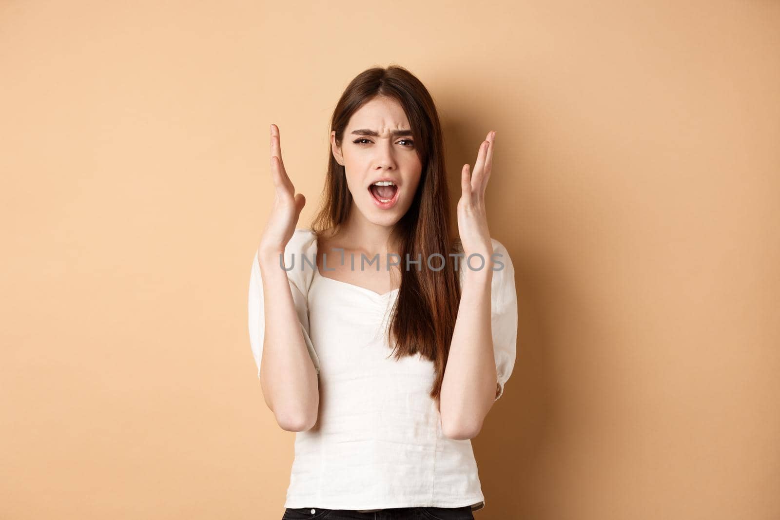 Frustarted woman shouting and shaking hands angry, looking mad, standing distressed on beige background.