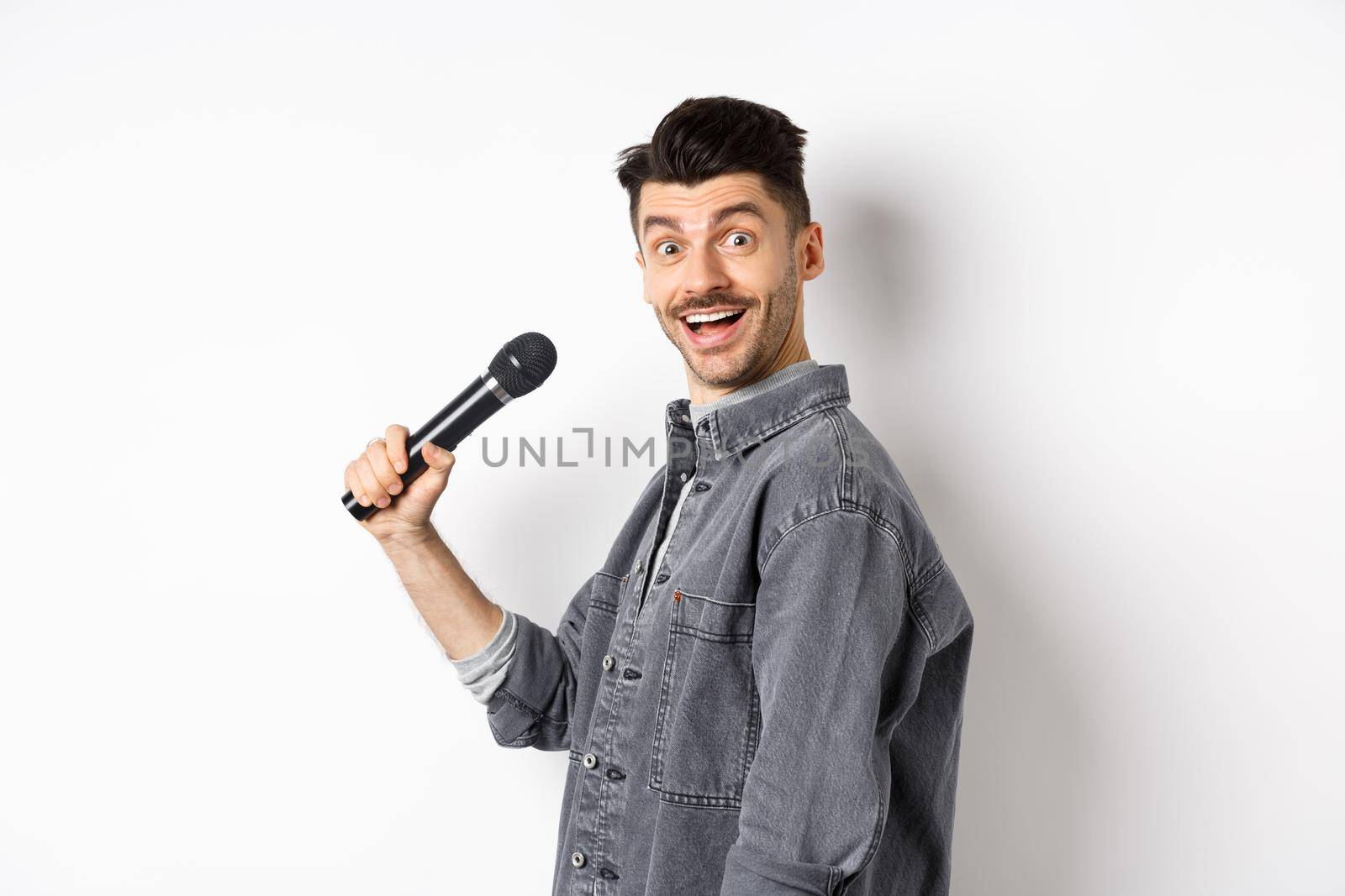 Profile of handsome smiling man holding mic, turn head at camera with excited face, singing karaoke and perform standup, standing on white background.