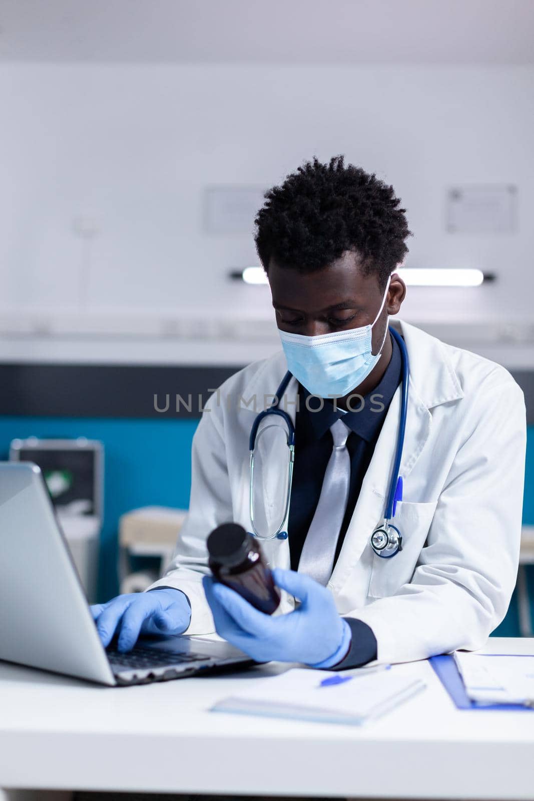 Black person with medic profession using laptop on desk holding bottle of medicament in medical cabinet. African american professional doctor typing prescription on keyboard while sitting