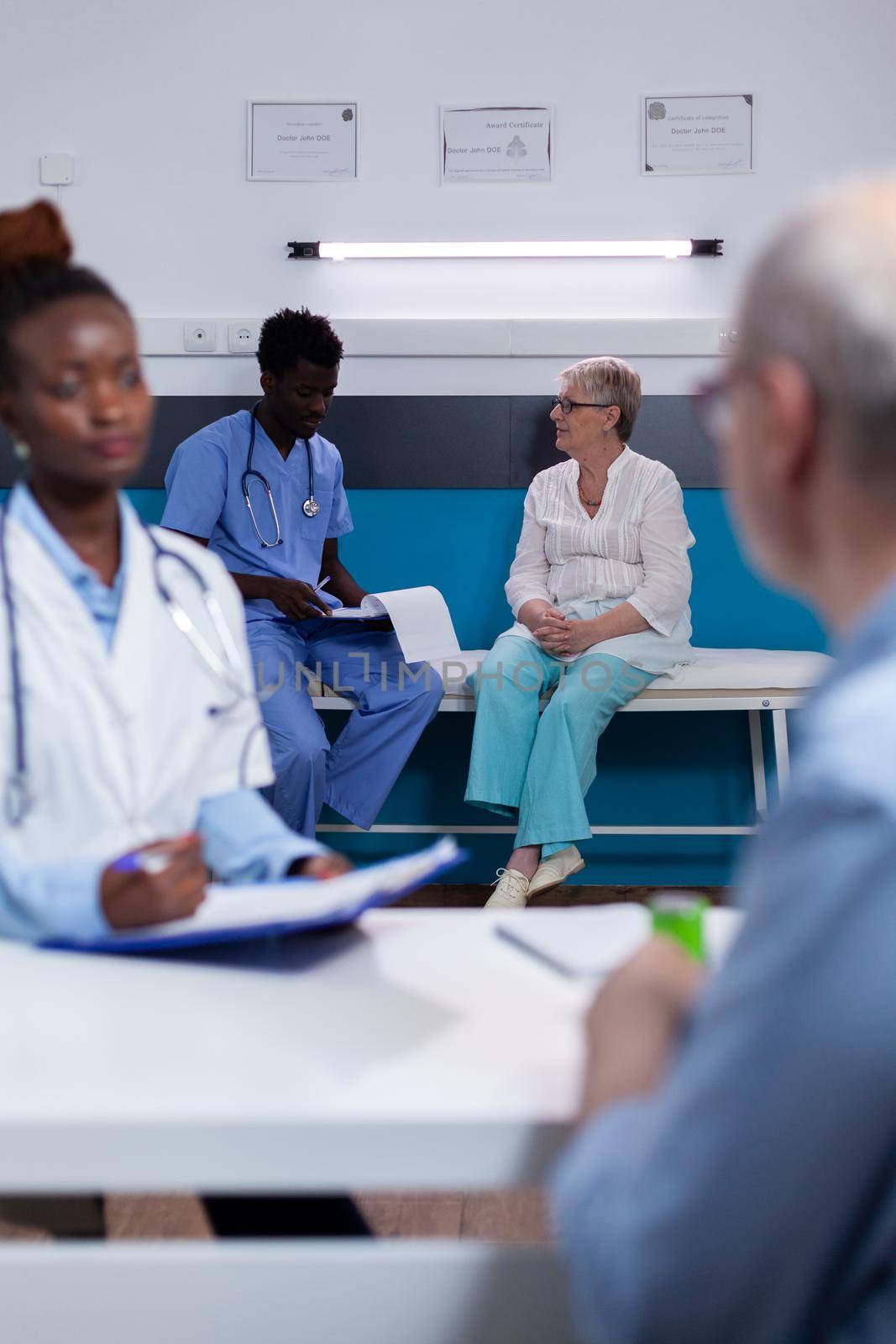 Black medic and caucasian old patient talking sitting at desk in medical cabinet. African american man with nurse profession consulting elder woman on bed in background at clinic