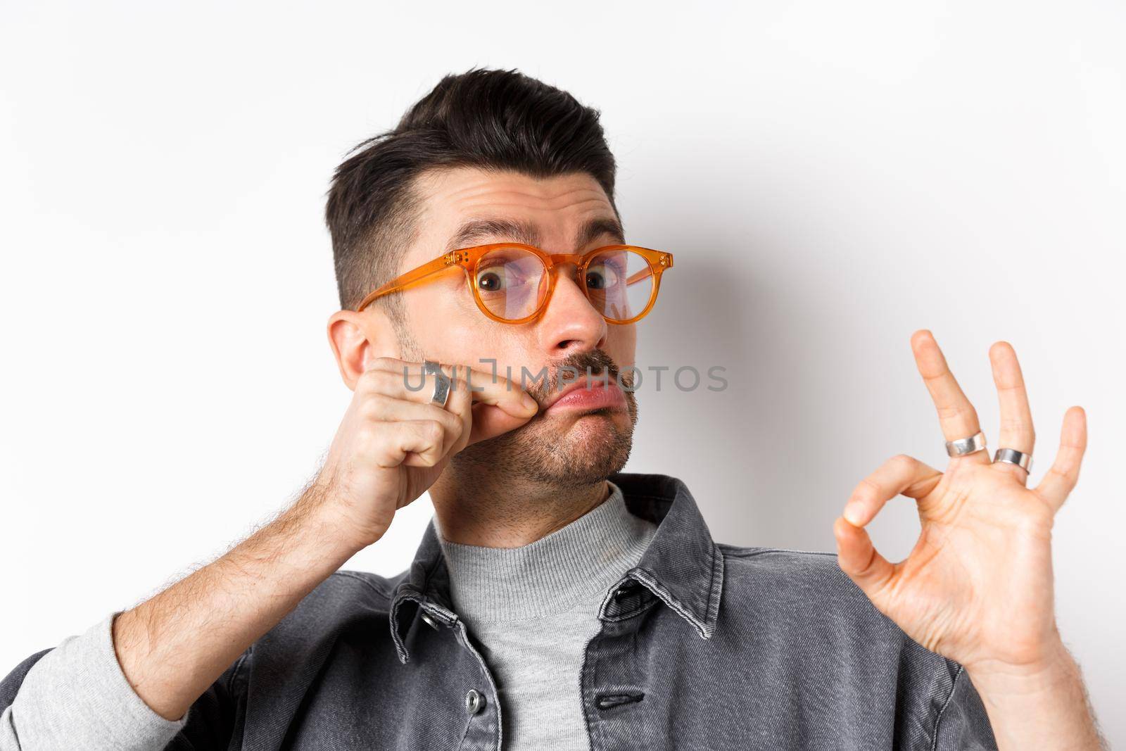 Funny young man in glasses touching his french moustache and showing okay, not bad sign, approve and like good product, standing on white background.