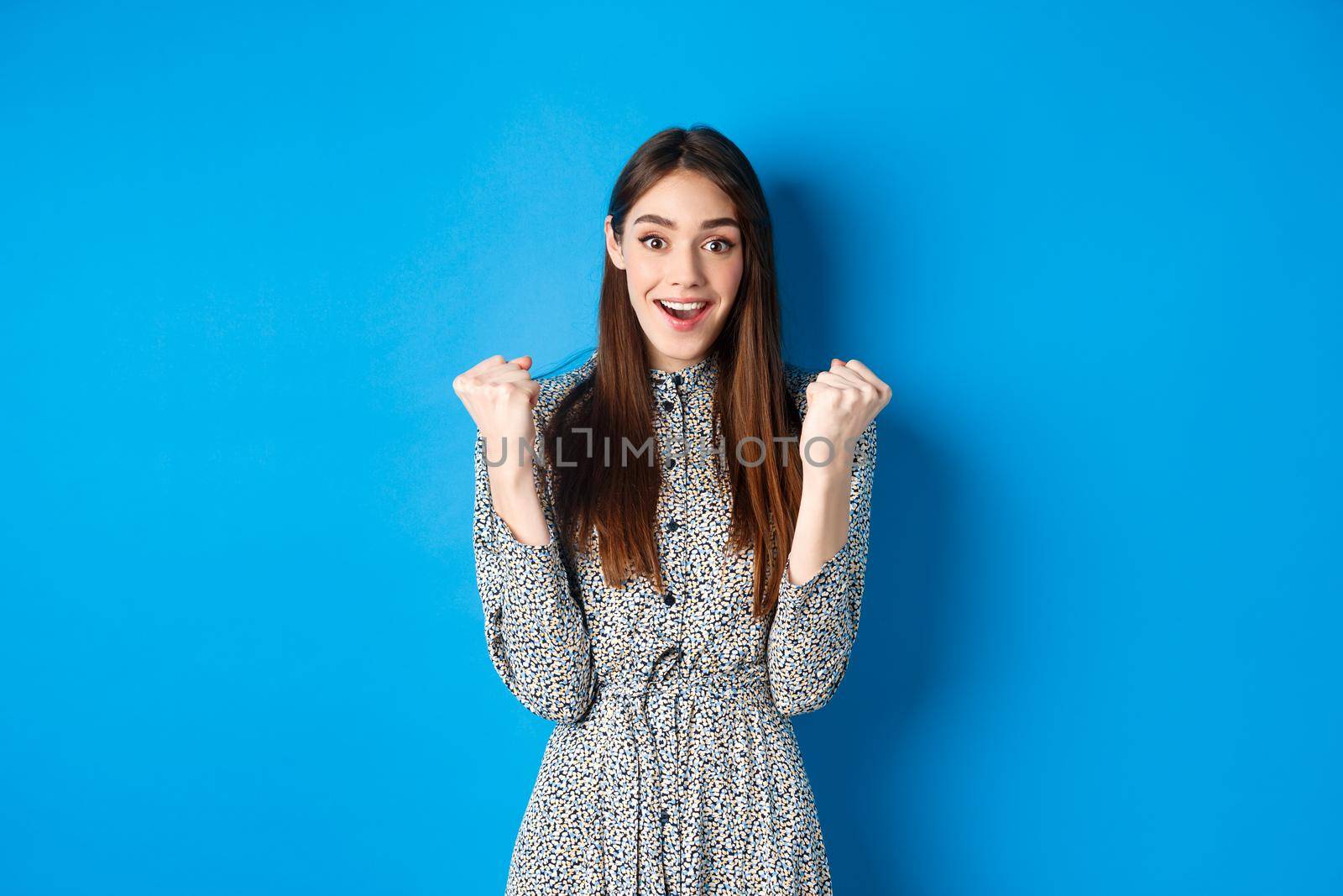 Excited woman looking relieved and hopeful, making fist pump and smiling, winning prize, celebrating achievement or victory, standing on blue background.