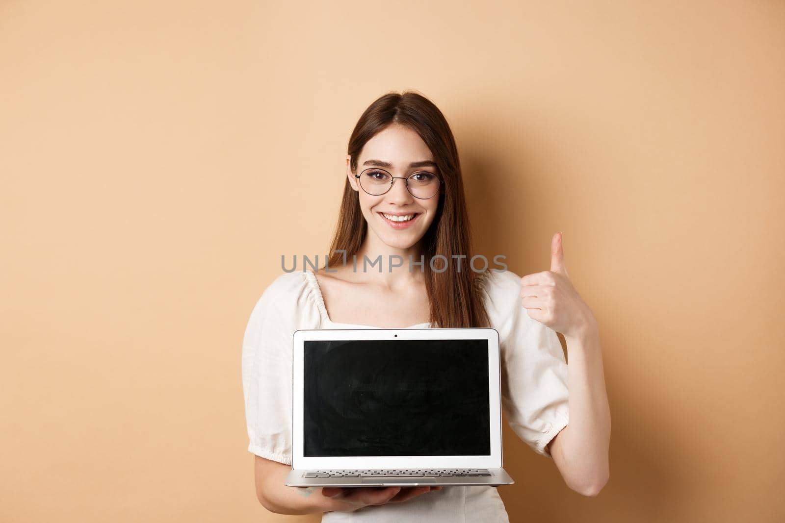 Smiling young woman in glasses showing laptop screen deal and thumb up, recommending offer, standing on beige background.