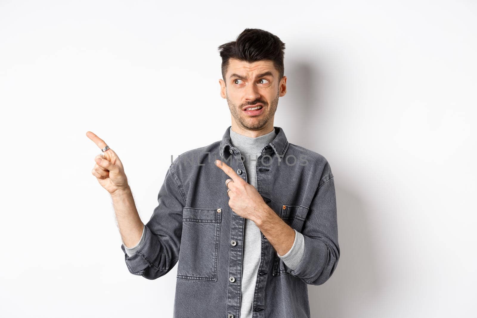 Skeptical and doubtful young man pointing fingers left at logo, look with dislike and confusion, standing on white background.