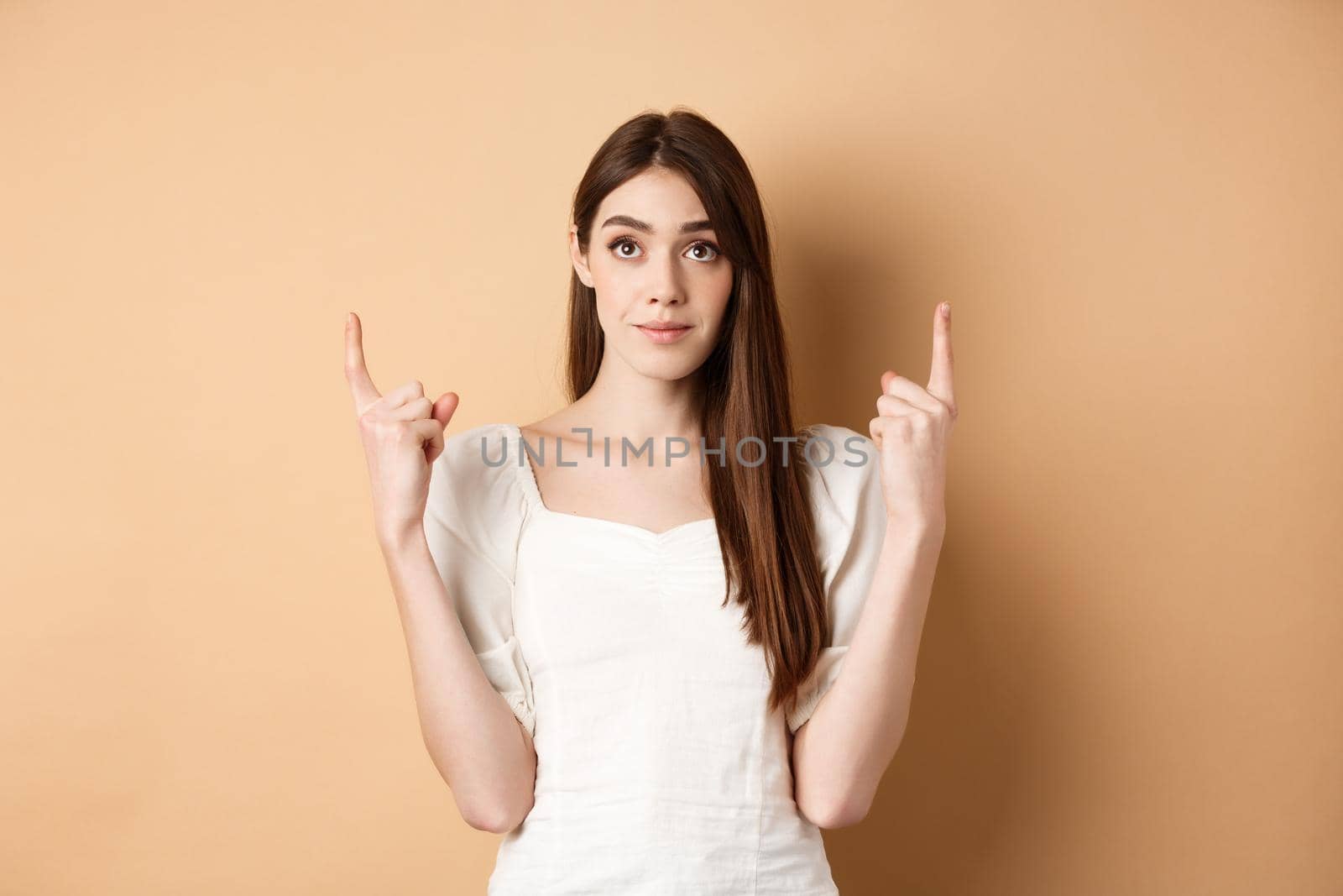 Hopeful romantic girl in white dress pointing fingers up, showing empty space for company logo, standing on beige background.