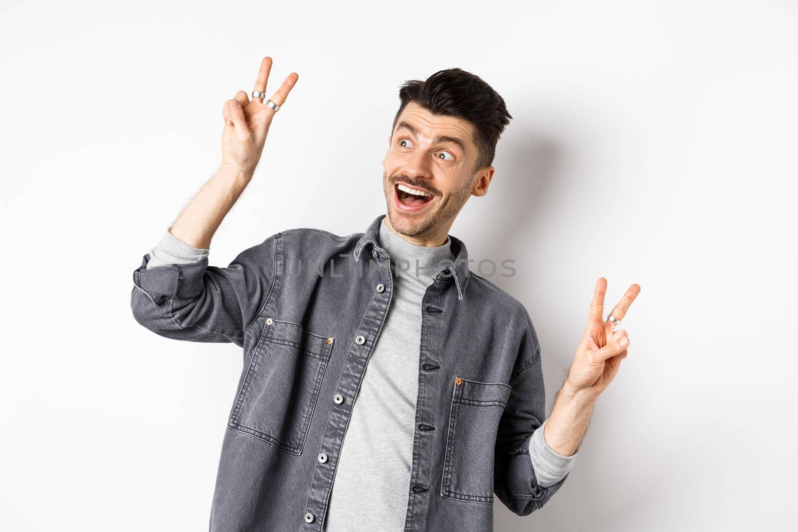 Funny young man showing v-signs and smiling at left side, looking at empty space with cheerful face expression, standing on white background.