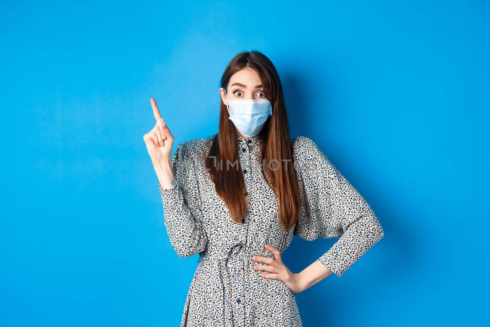 Healthy people and covid-19 pandemic concept. Excited woman in medical mask pointing at upper left corner, standing on blue background.