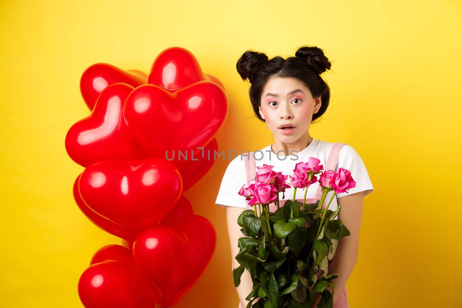 Happy Valentines day. Surprised asian girl receive bouquet of pink roses from lover, looking with awe at camera, standing near red heart balloons, yellow background.