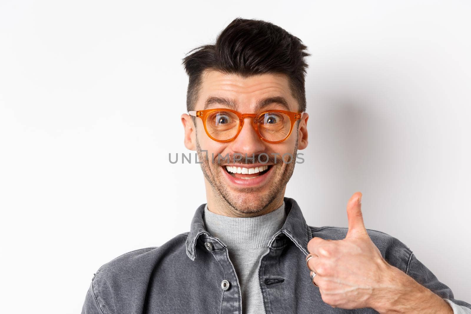 Close up portrait of excited funny guy in eyewear showing thumbs up, smiling supportive, praise and recommend cool store, standing on white background.